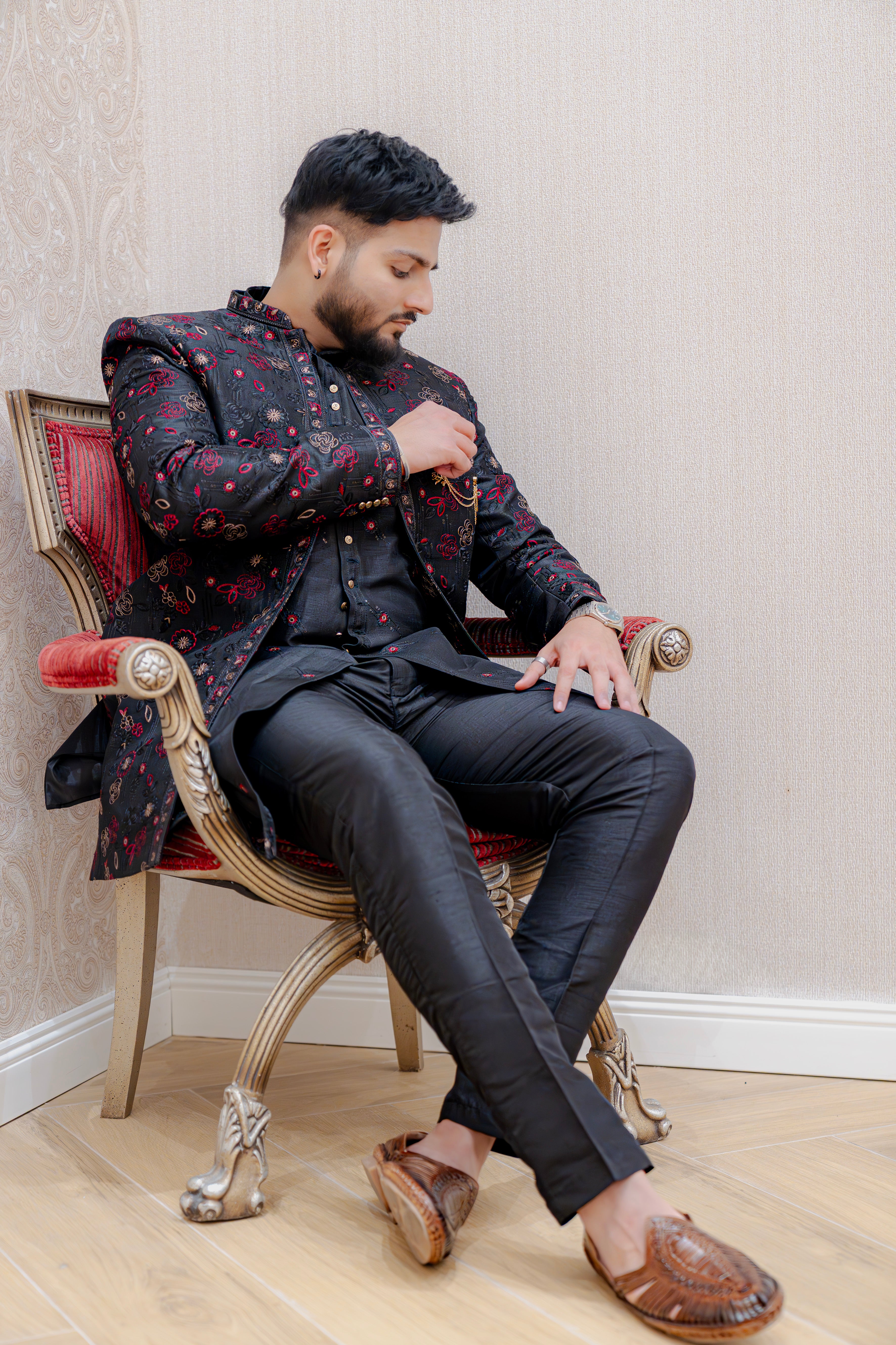 Man seated on an ornate chair wearing a black Indo-Western sherwani jacket with red shimmer floral jacquard over a black kurta and trousers, accented with a red pocket square, against a light wallpapered background.