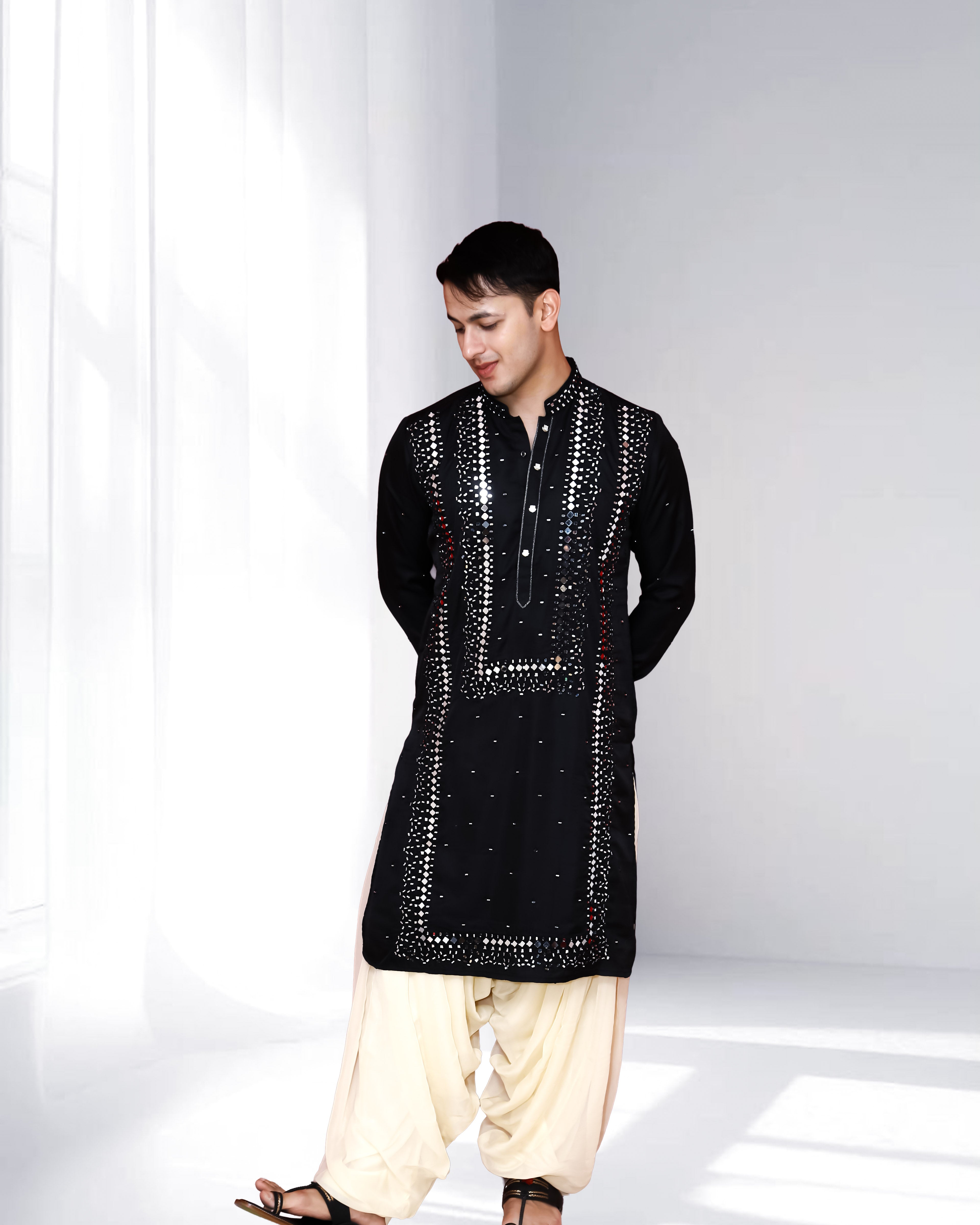 Man wearing a black mirrorwork embroidered kurta paired with a cream dhoti, styled in a modern minimal indoor setting.
