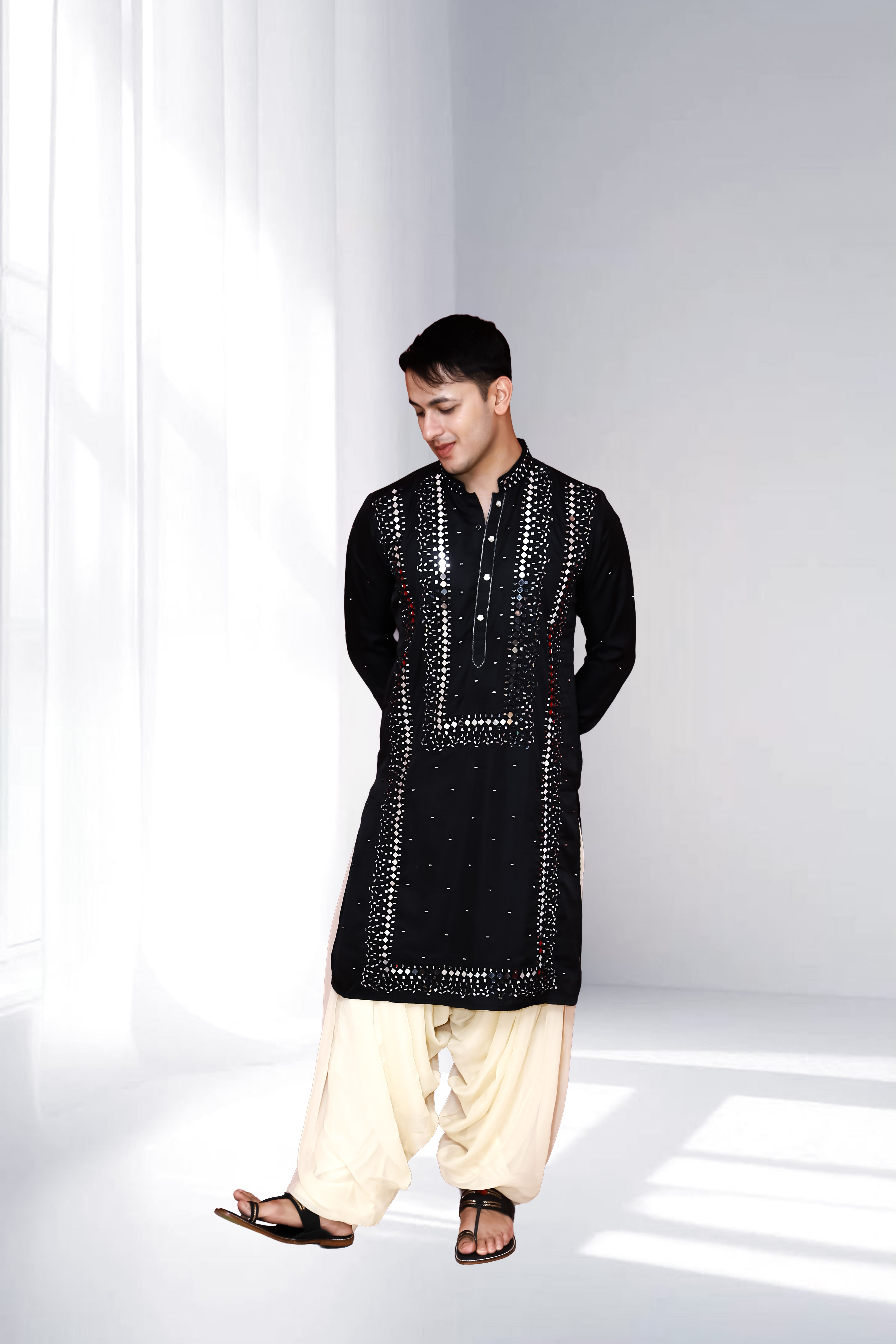 Man wearing a black mirrorwork embroidered kurta paired with a cream dhoti, styled in a modern minimal indoor setting.