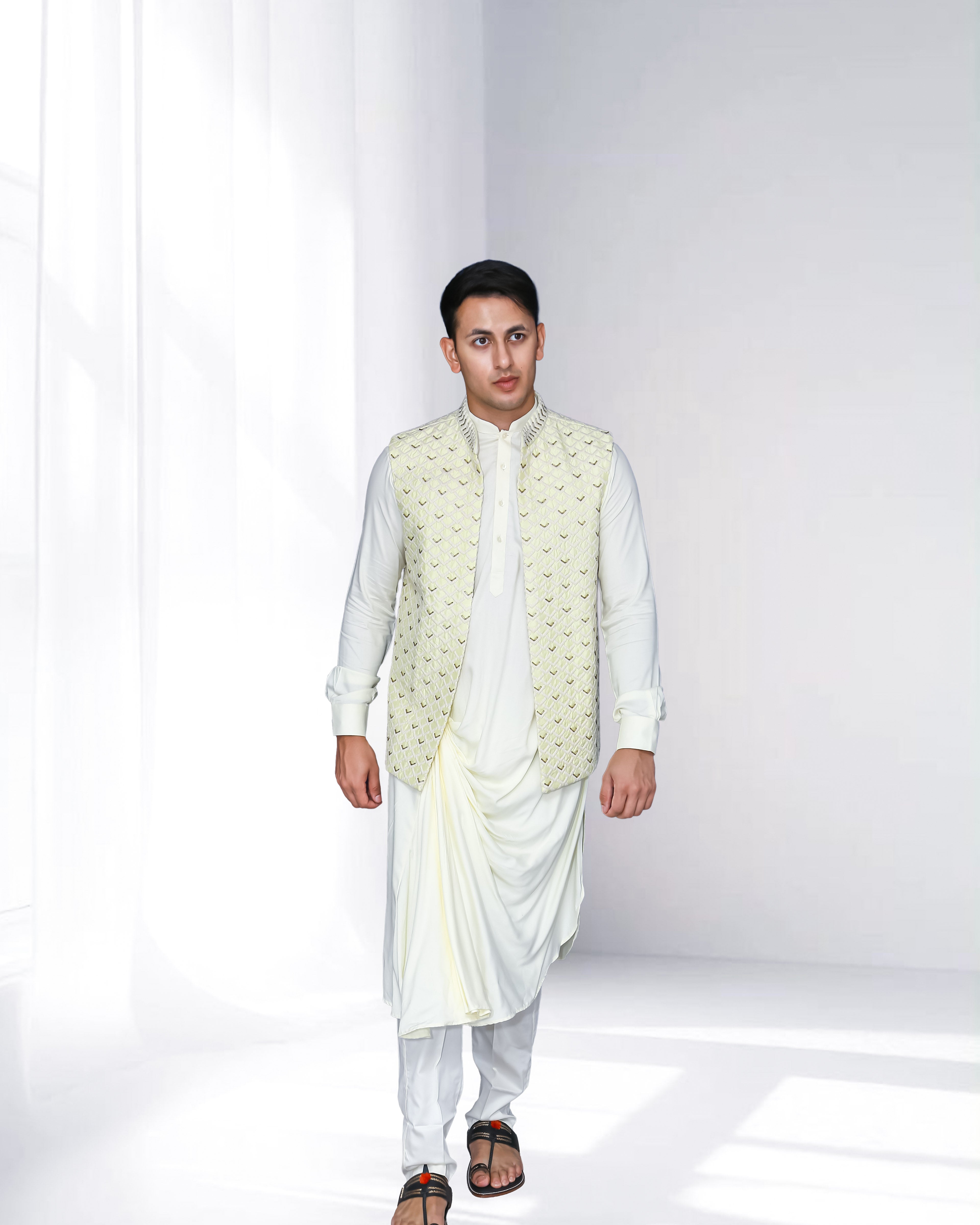 Man in an ivory indo-western kurta with draped detailing, styled with a cream embroidered nehru jacket, standing against a soft-lit minimal background.