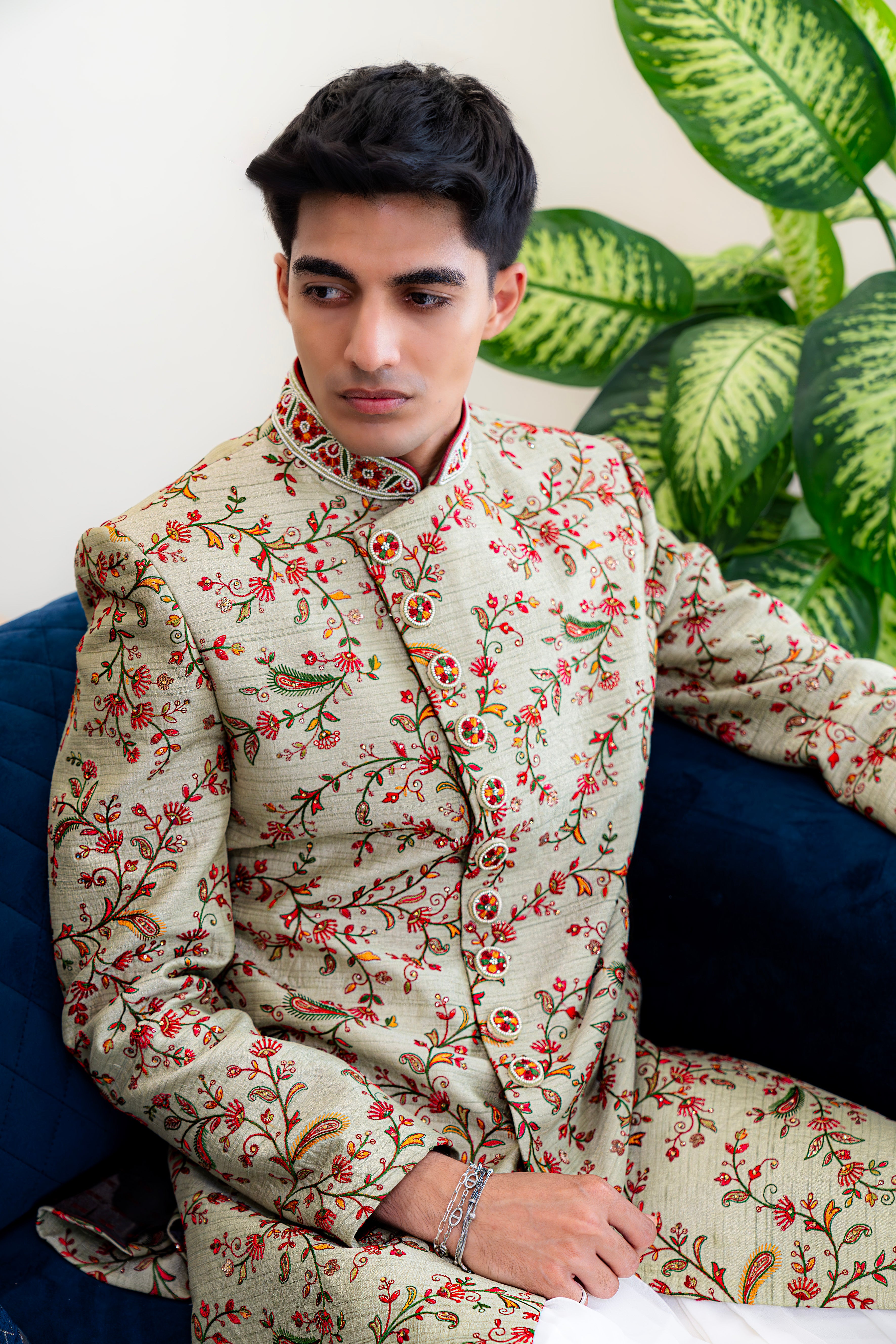 Man wearing a floral patterned suit sitting on a blue couch with green plants in the background