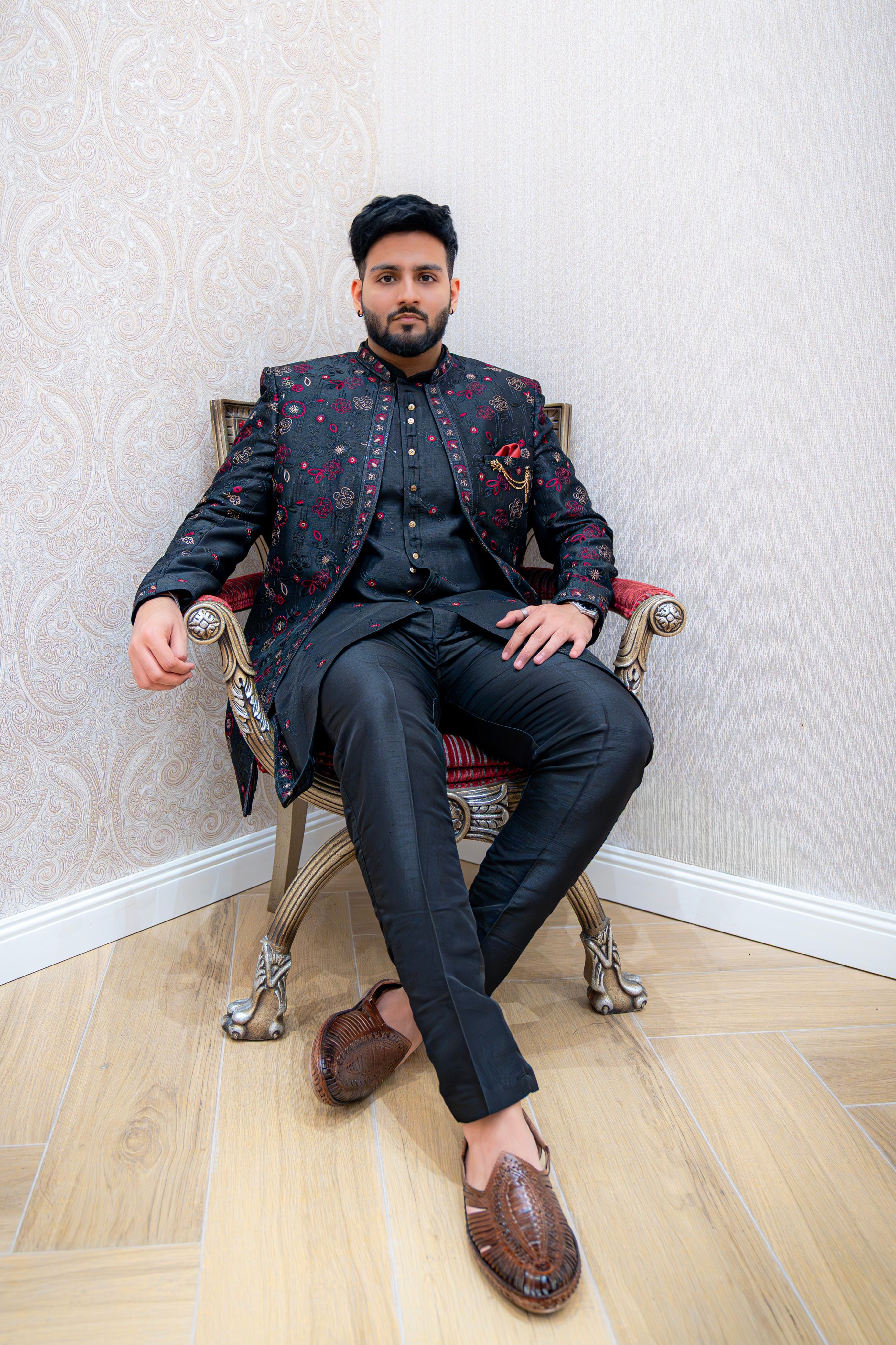 Man seated on an ornate chair wearing a black Indo-Western sherwani jacket with red shimmer floral jacquard over a black kurta and trousers, accented with a red pocket square, against a light wallpapered background.