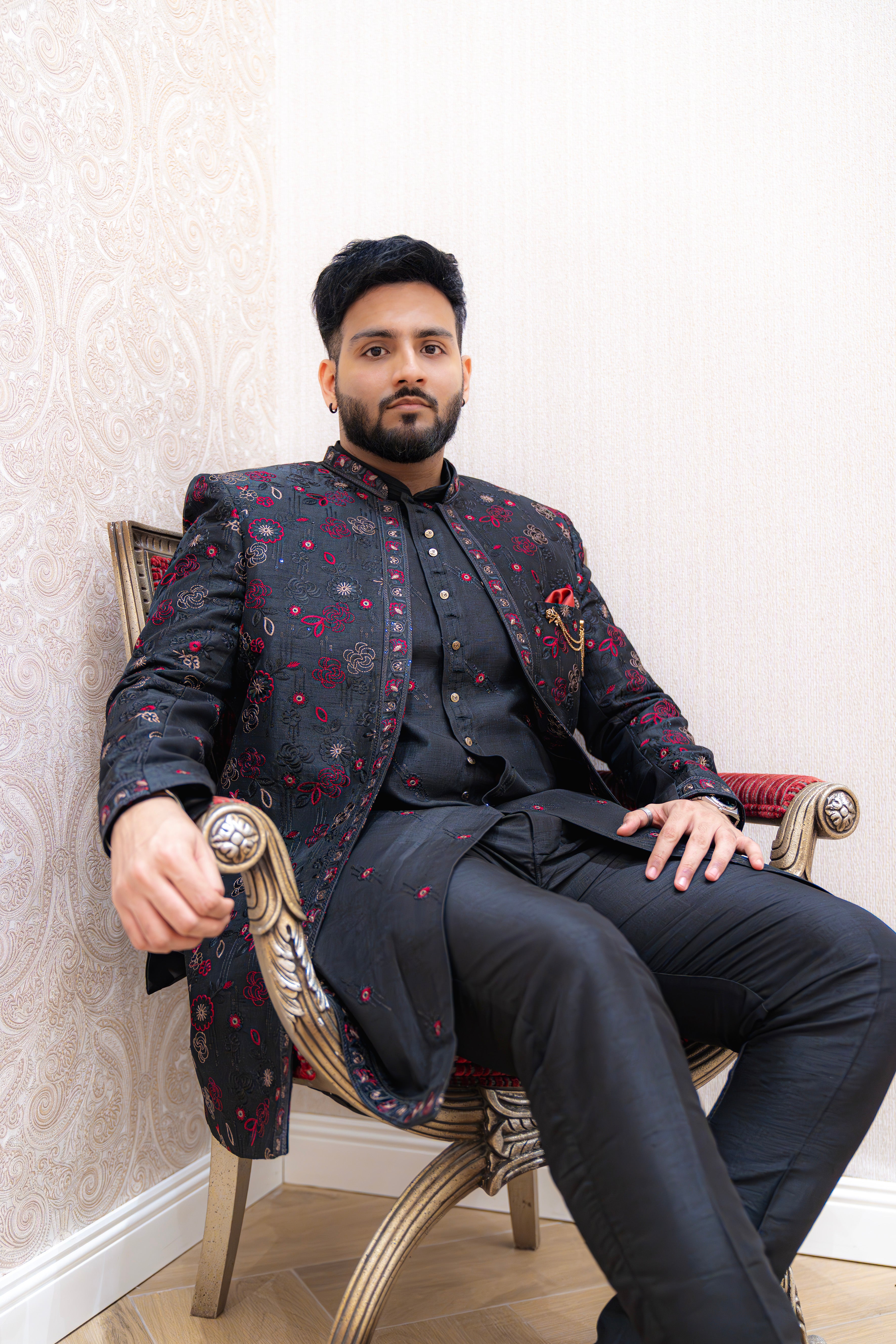 Man seated on an ornate chair wearing a black Indo-Western sherwani jacket with red shimmer floral jacquard over a black kurta and trousers, accented with a red pocket square, against a light wallpapered background.