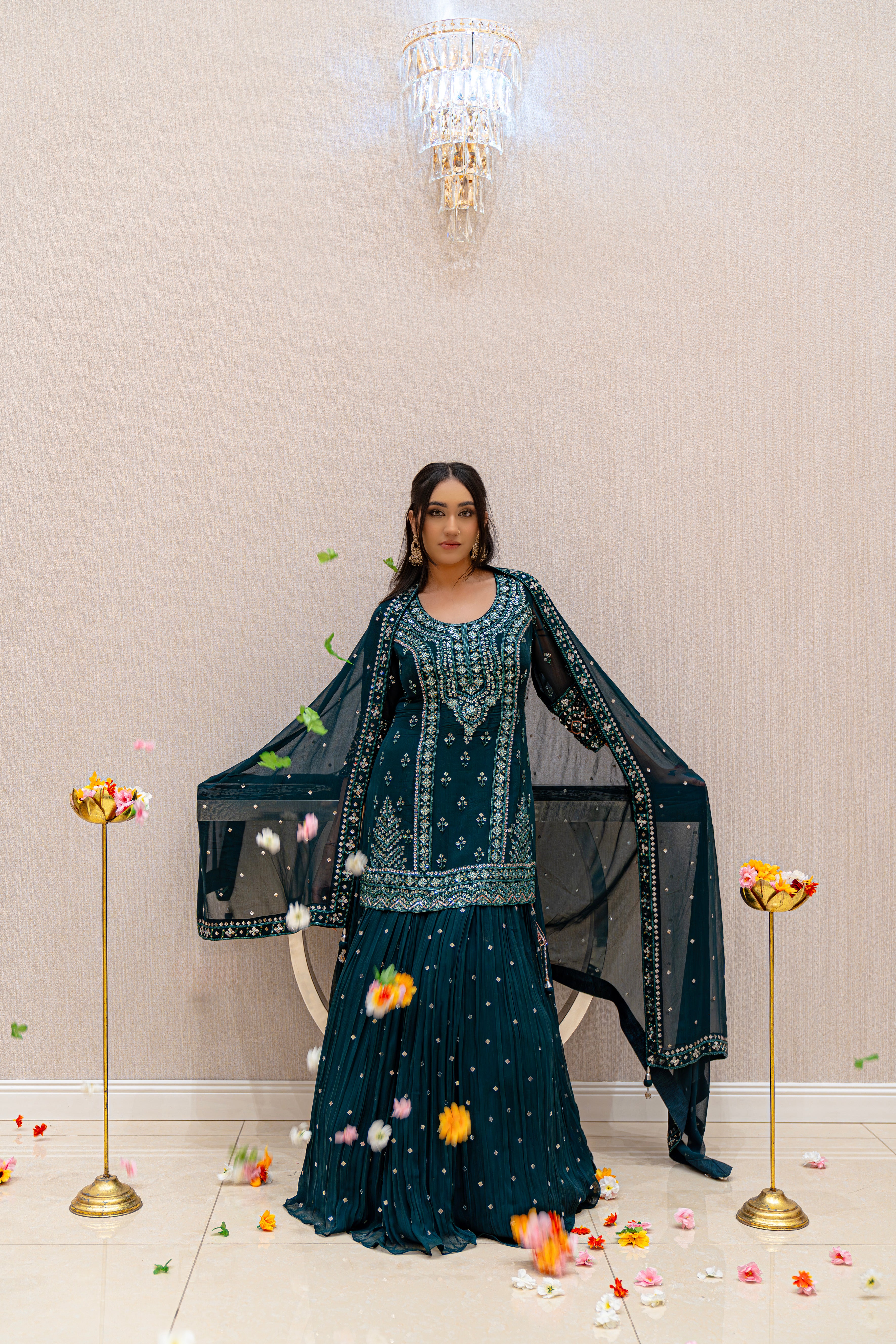 Woman in an emerald green embroidered outfit standing in a decorated room with flowers and a chandelier.