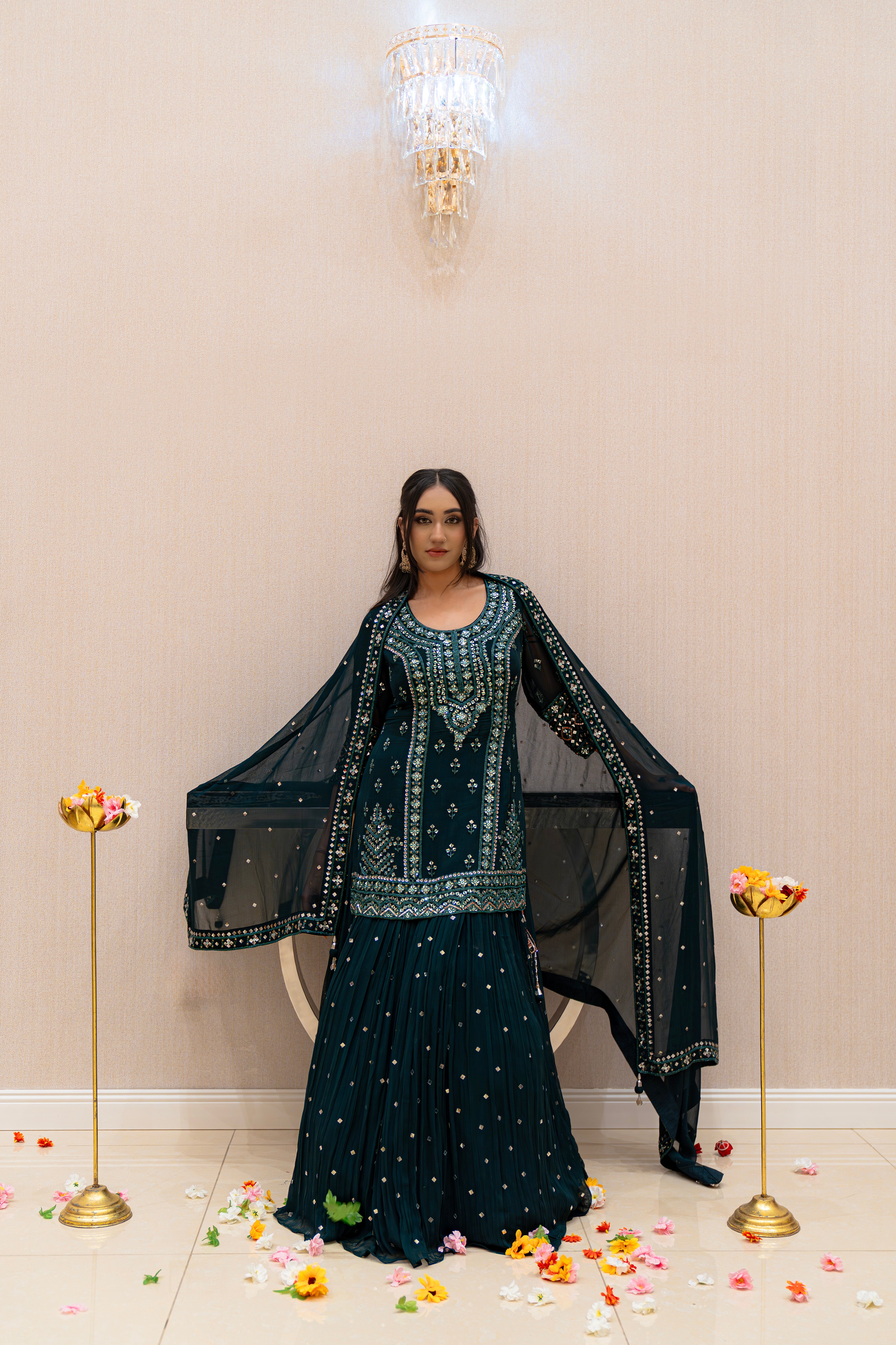 Woman in a green blue embroidered traditional outfit standing in a decorated room with flowers on the floor.