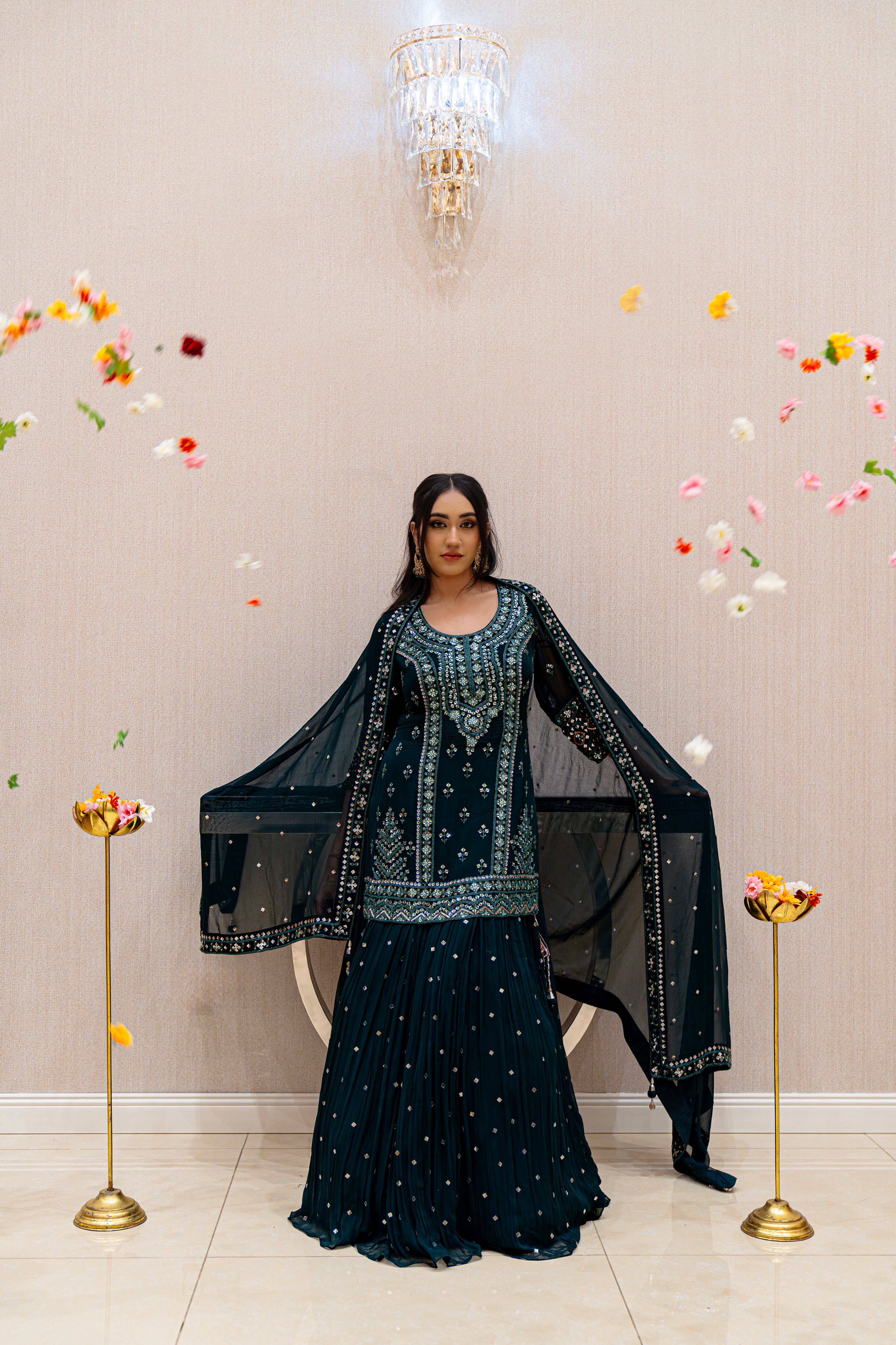 Woman in an emerald embroidered traditional outfit standing against a beige wall with decorative elements.