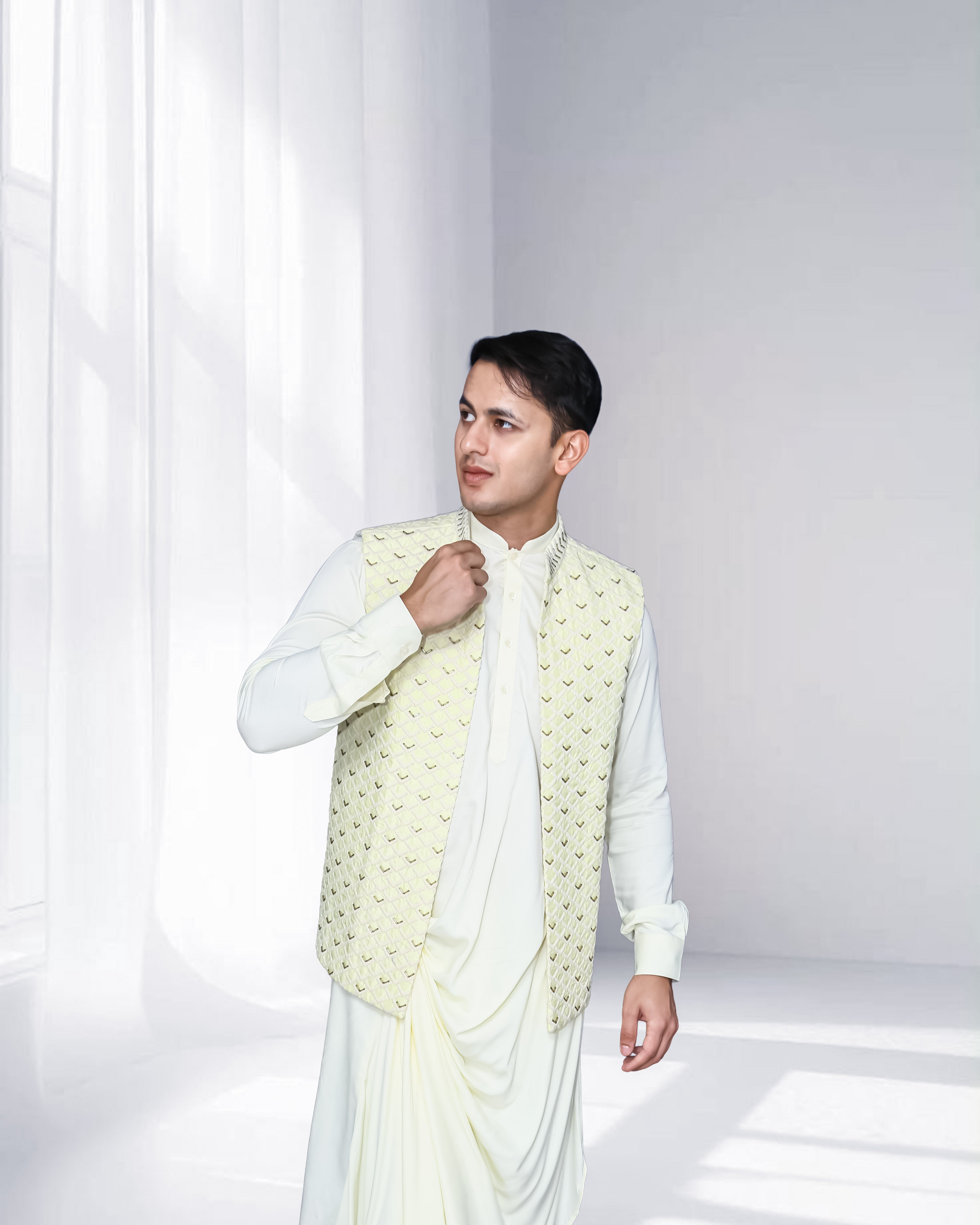 Man in an ivory indo-western kurta with draped detailing, styled with a cream embroidered nehru jacket, standing against a soft-lit minimal background.