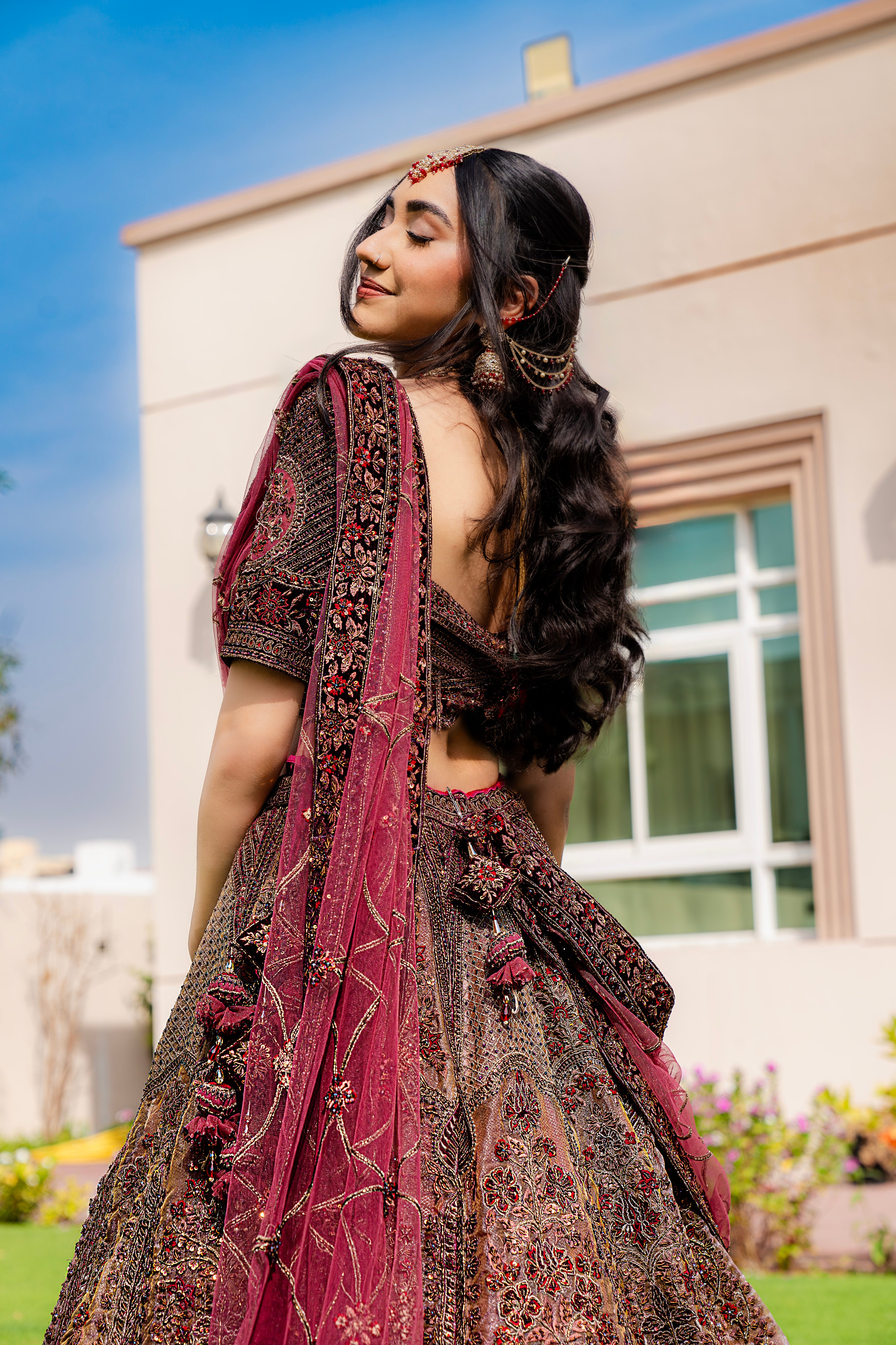 Woman wearing a maroon and brown embroidered bridal lehenga with matching dupatta, standing outdoors, ideal for weddings and receptions.