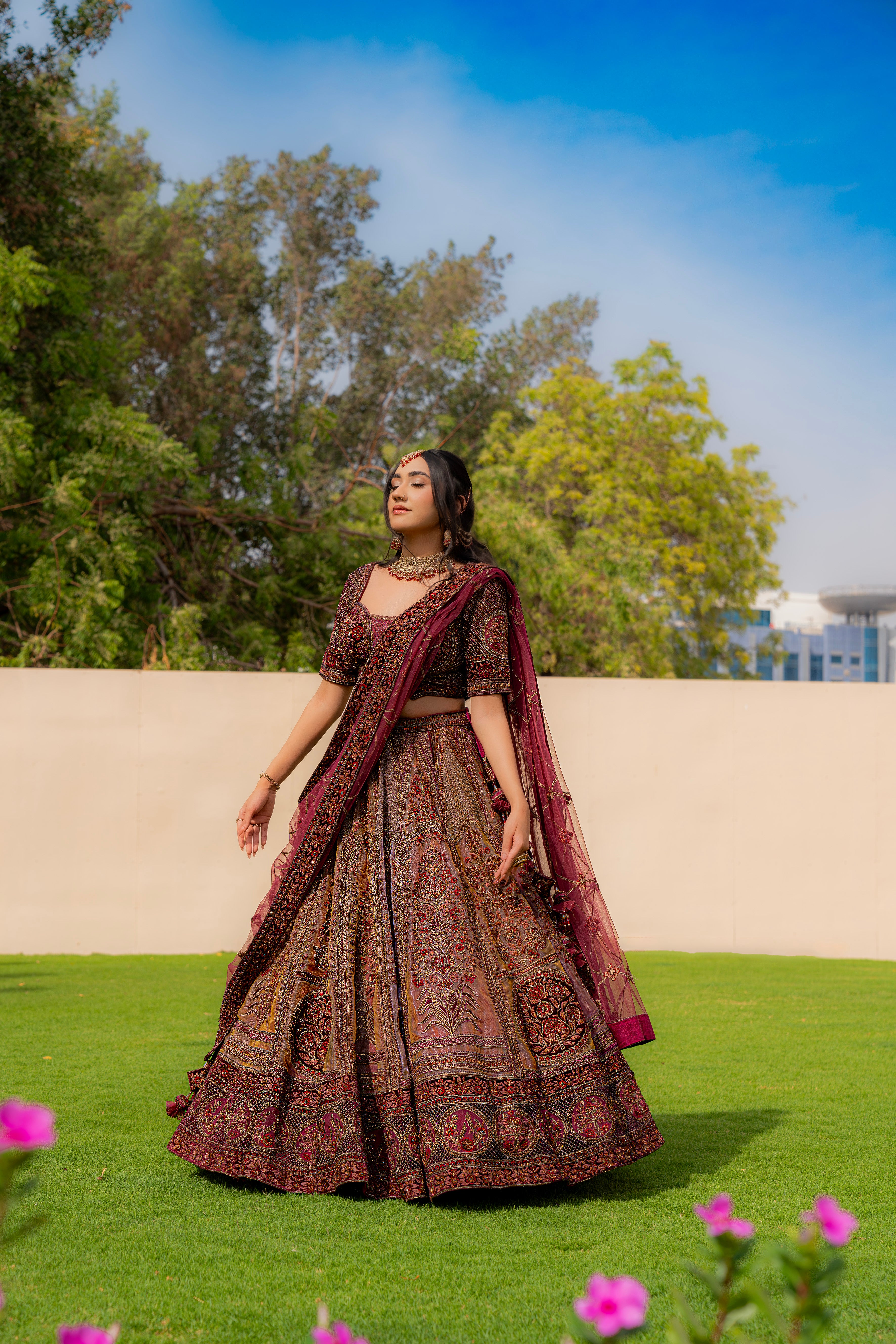 Woman wearing a maroon and brown embroidered bridal lehenga with matching dupatta, standing outdoors, ideal for weddings and receptions.