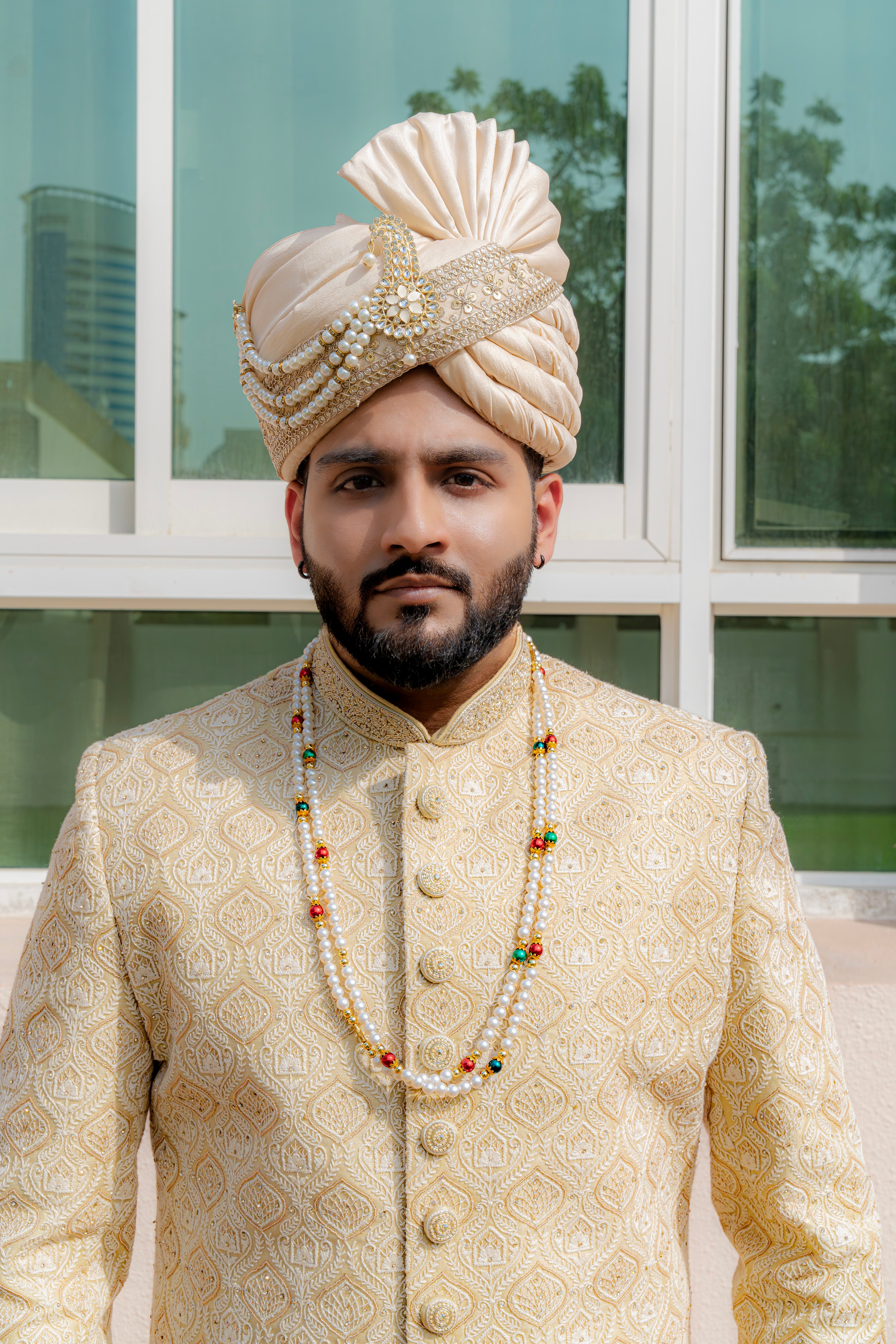 Man in traditional pale yellow sherwani standing outdoors with a window and plants in the background