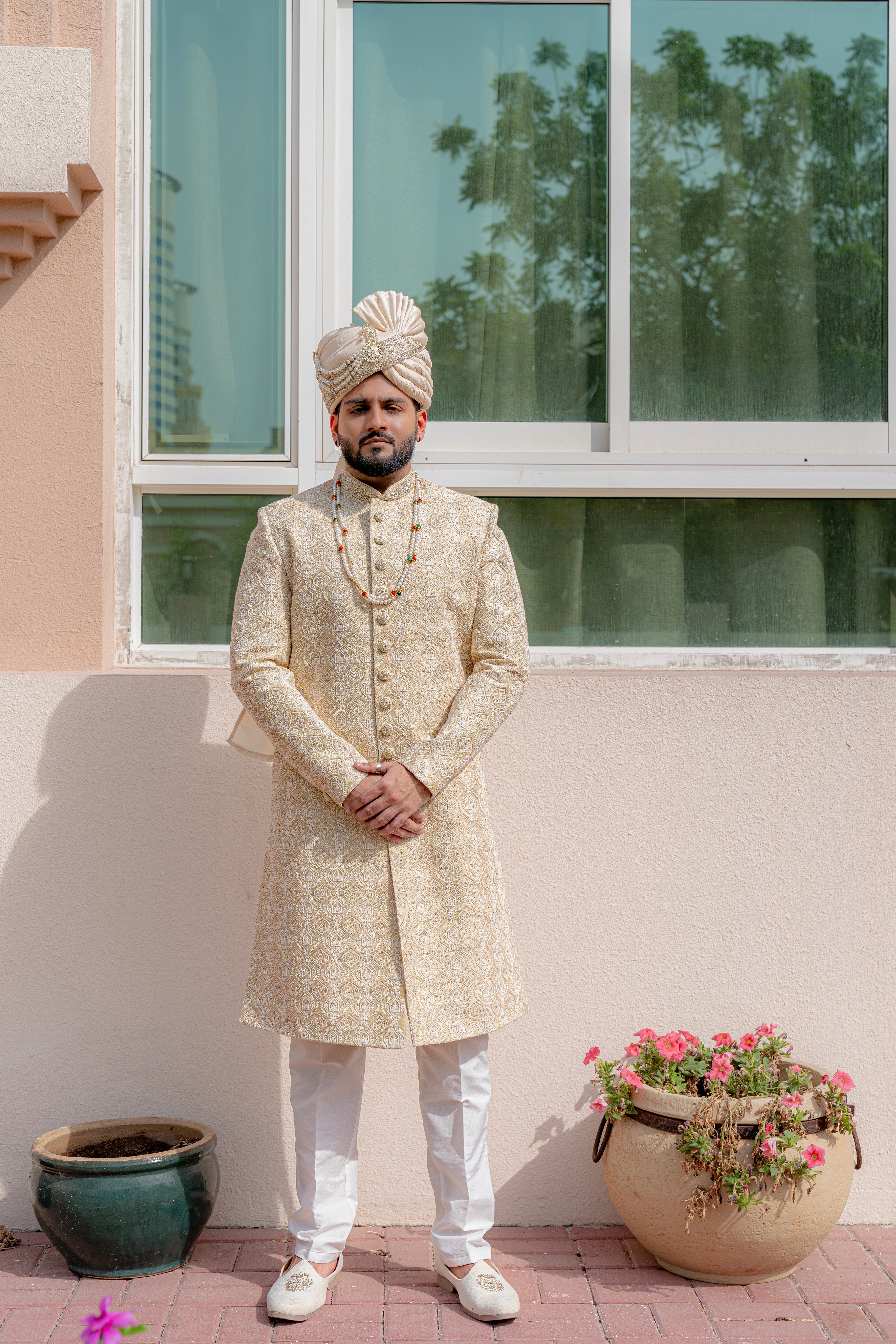 Man in traditional pale yellow sherwani standing outdoors with a window and plants in the background
