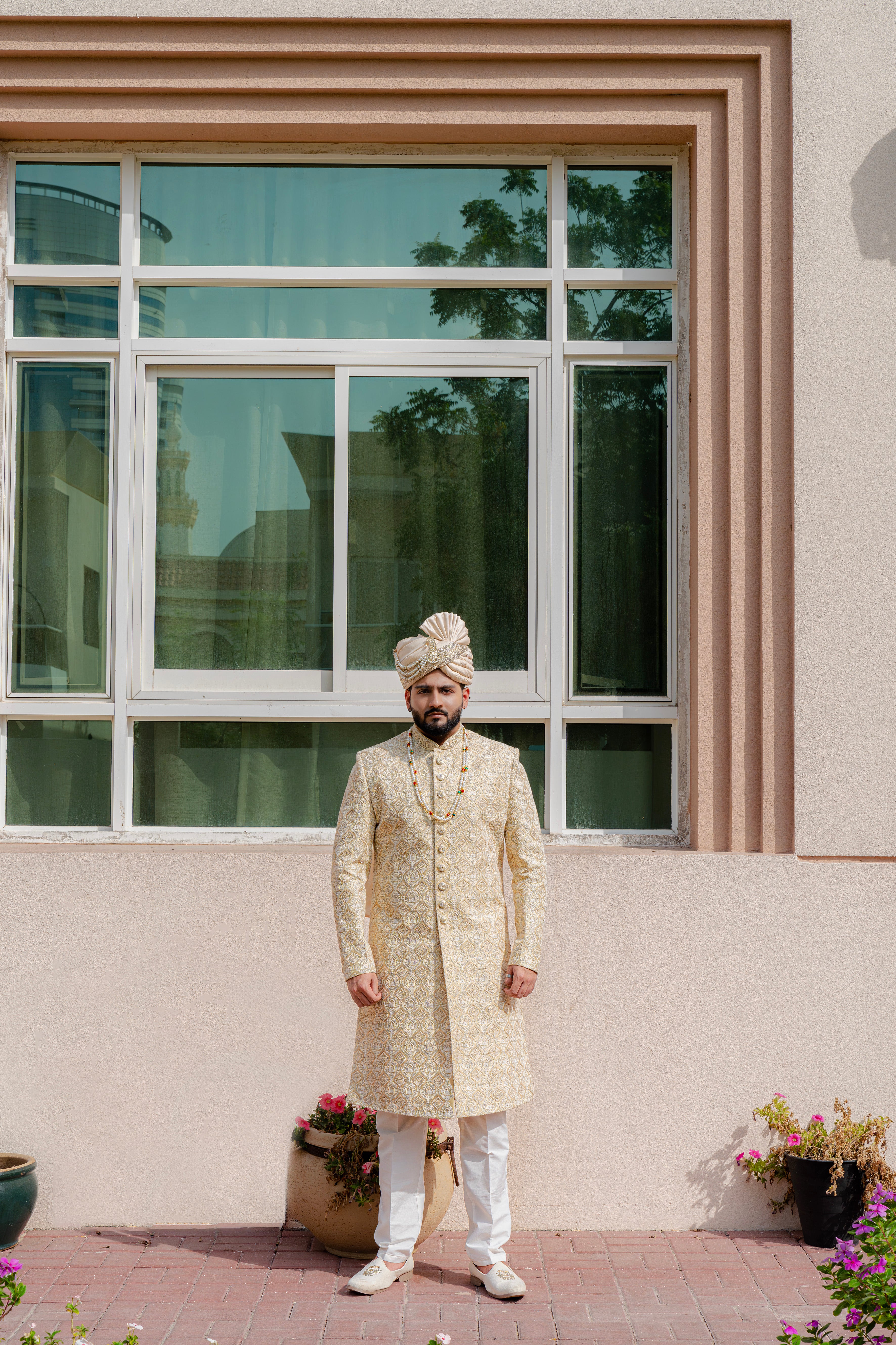 Man in traditional cream sherwani standing in front of a window with decorative plants.