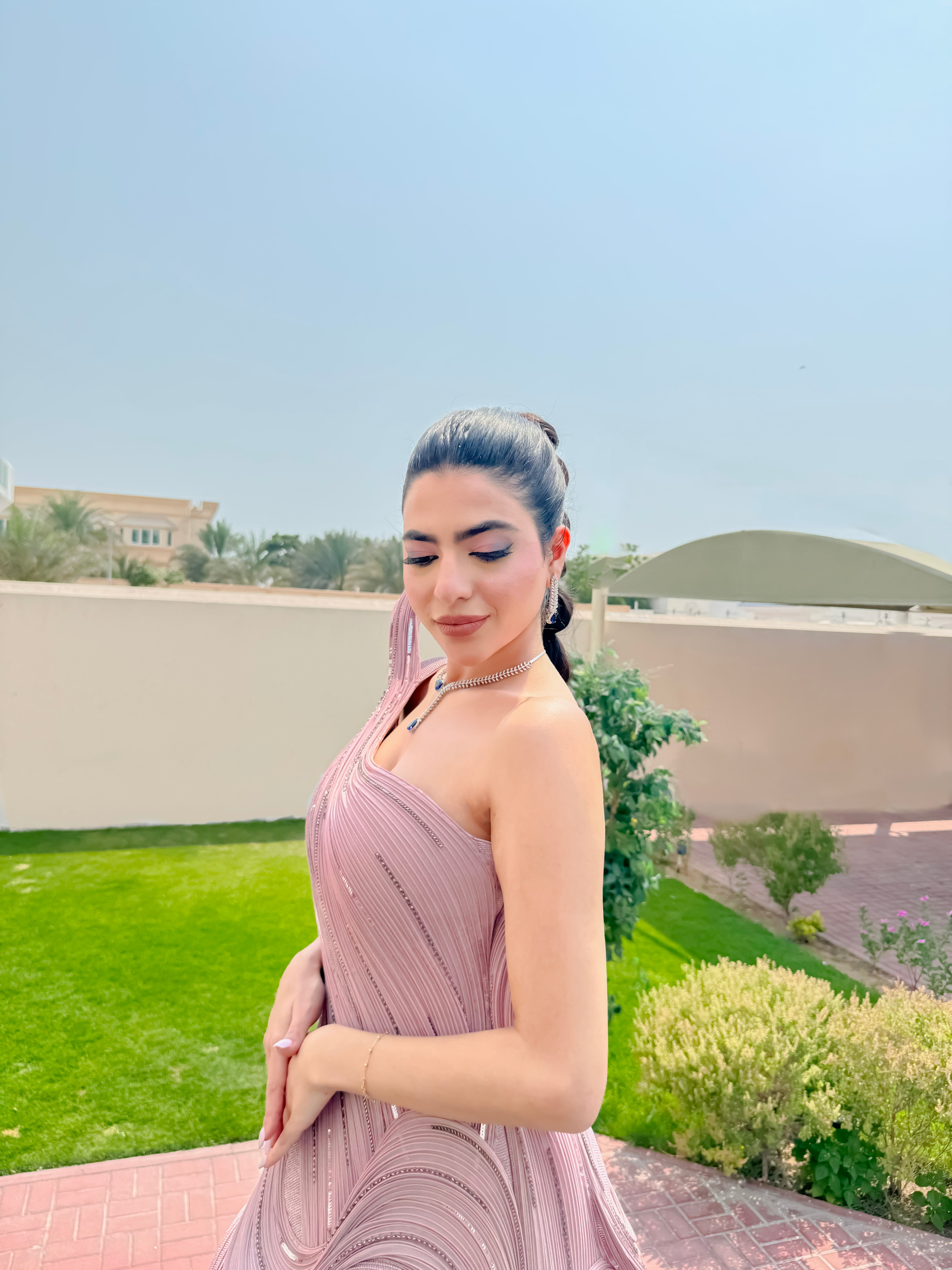 Woman in a pink dress standing outdoors with greenery and a clear sky in the background