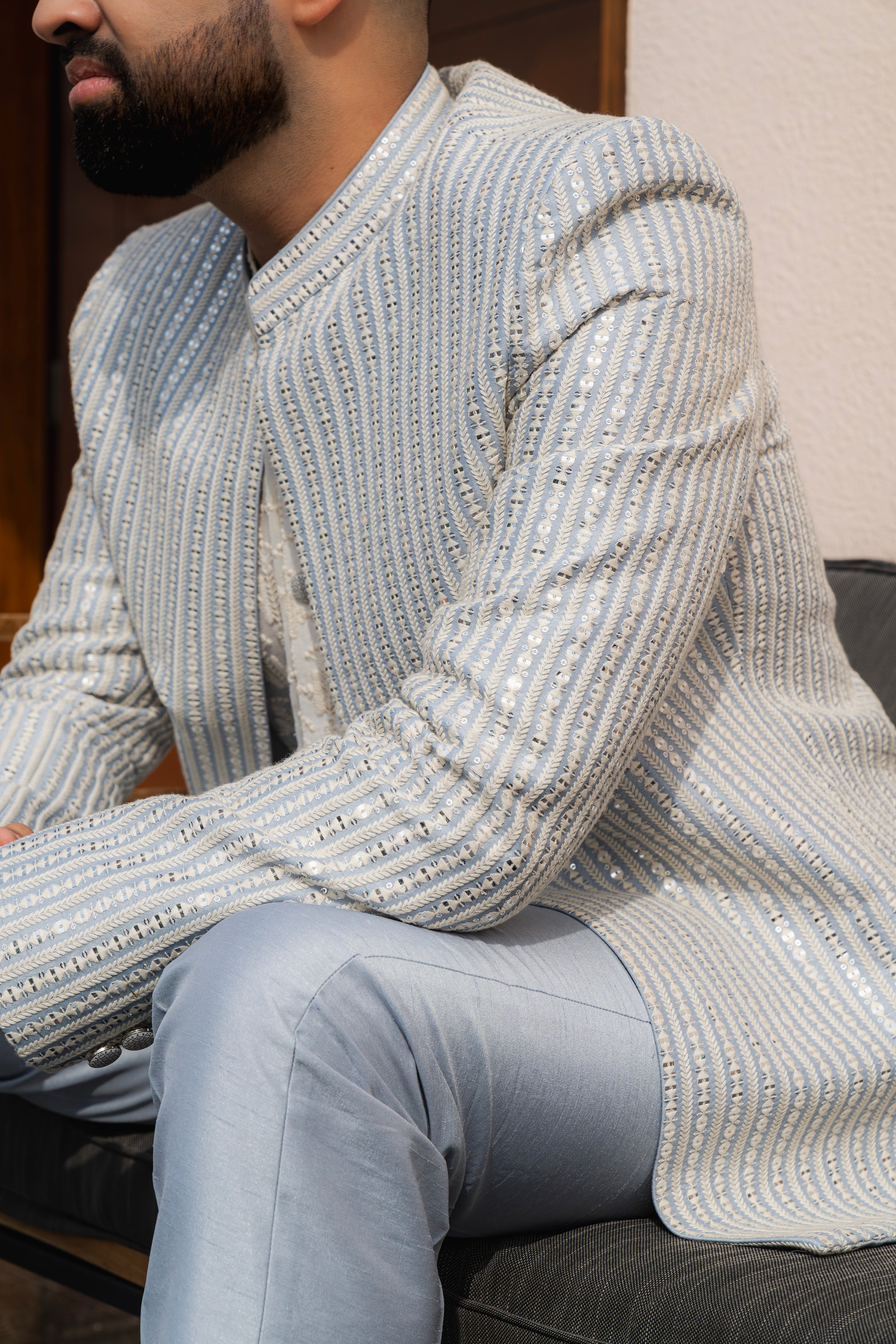 Man wearing a silver textured bandhgala jacket with subtle embroidery, sitting outdoors, ideal for weddings and festive occasions.