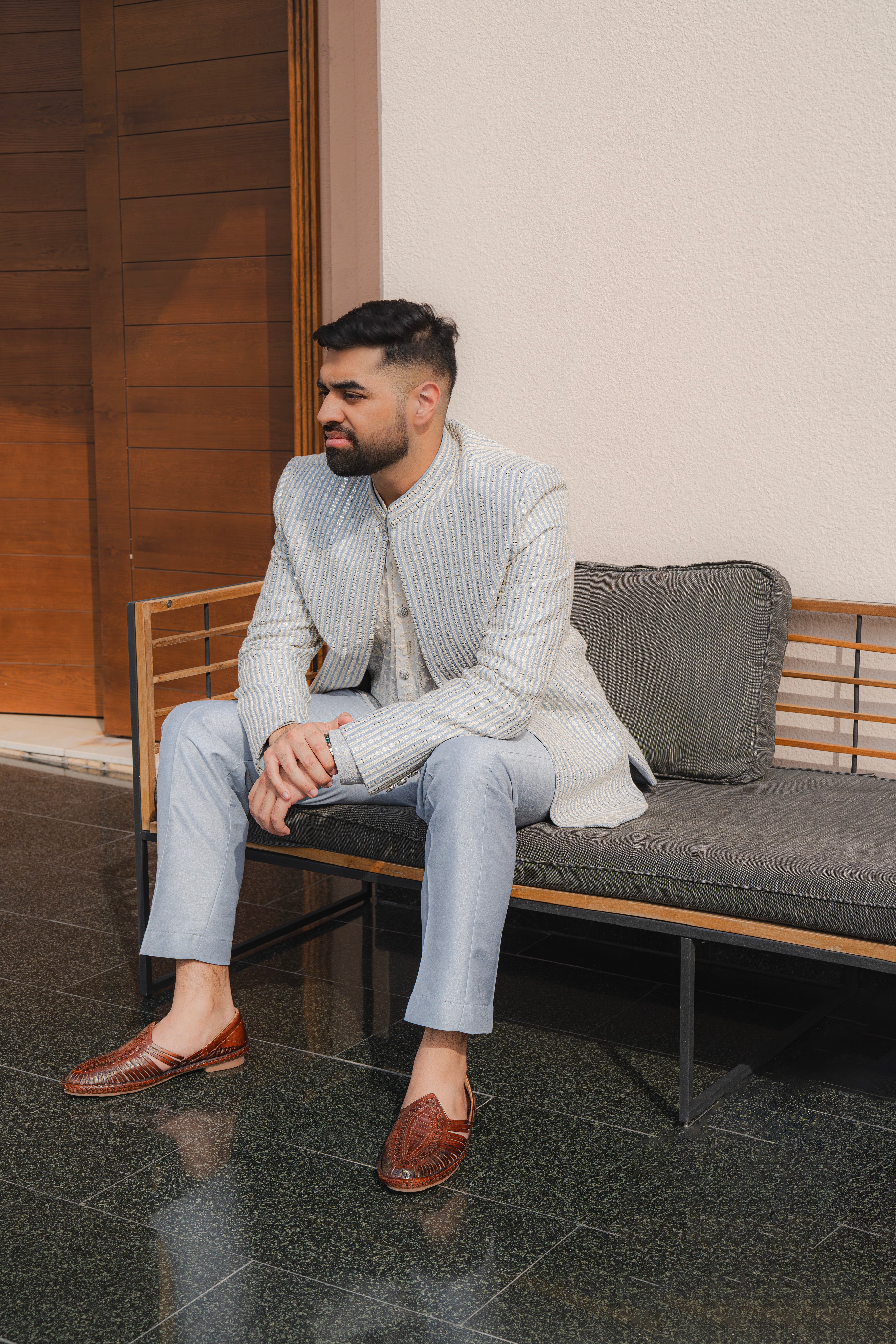 Man wearing a baby blue and silver textured bandhgala jacket with subtle embroidery, sitting outdoors, ideal for weddings and festive occasions.
