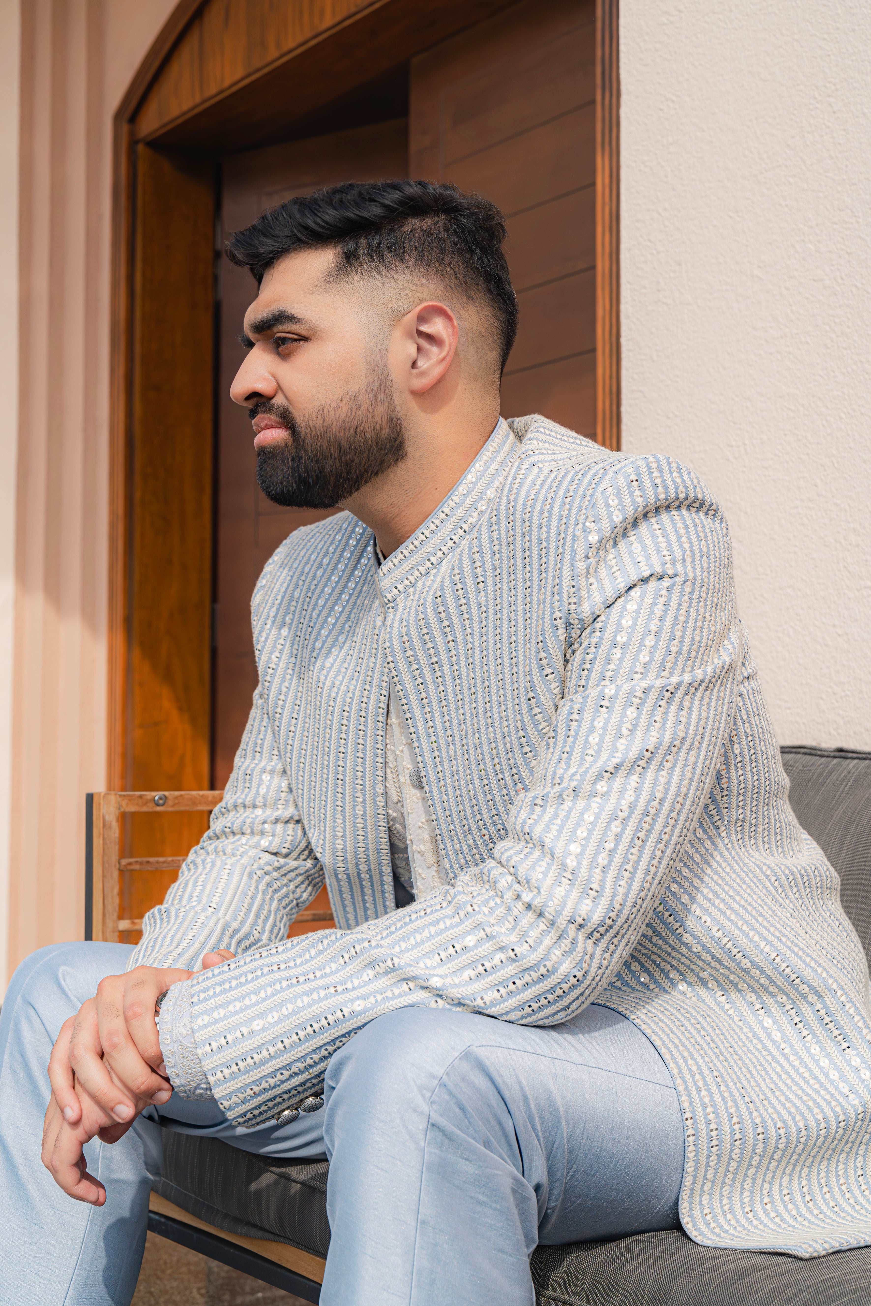 Man wearing a baby blue and silver textured bandhgala jacket with subtle embroidery, sitting outdoors, ideal for weddings and festive occasions.