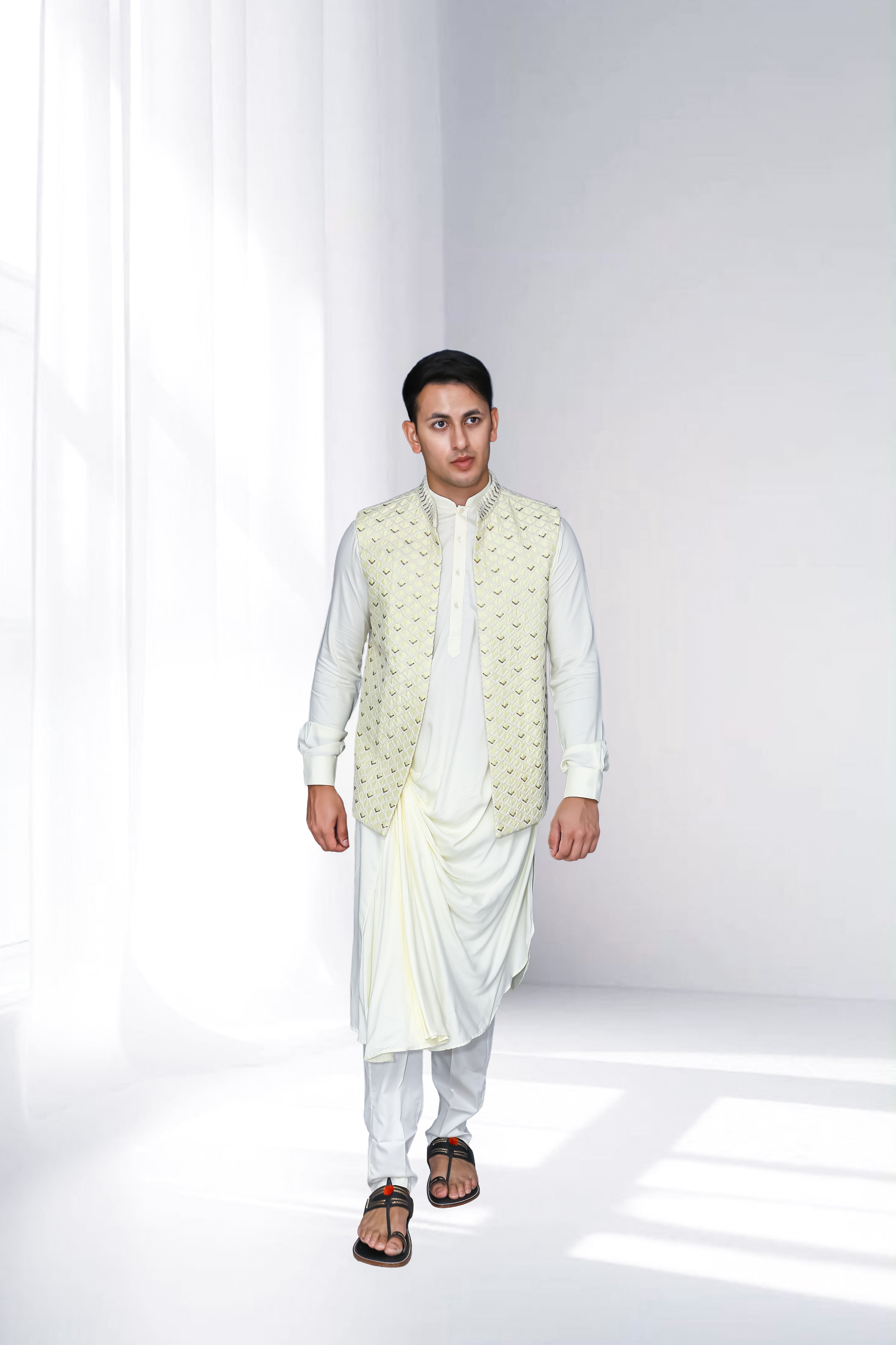 Man in an ivory indo-western kurta with draped detailing, styled with a cream embroidered nehru jacket, standing against a soft-lit minimal background.