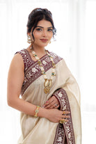 Woman wearing an elegant cream saree with a maroon embroidered border, styled indoors, perfect for weddings and festive occasions.