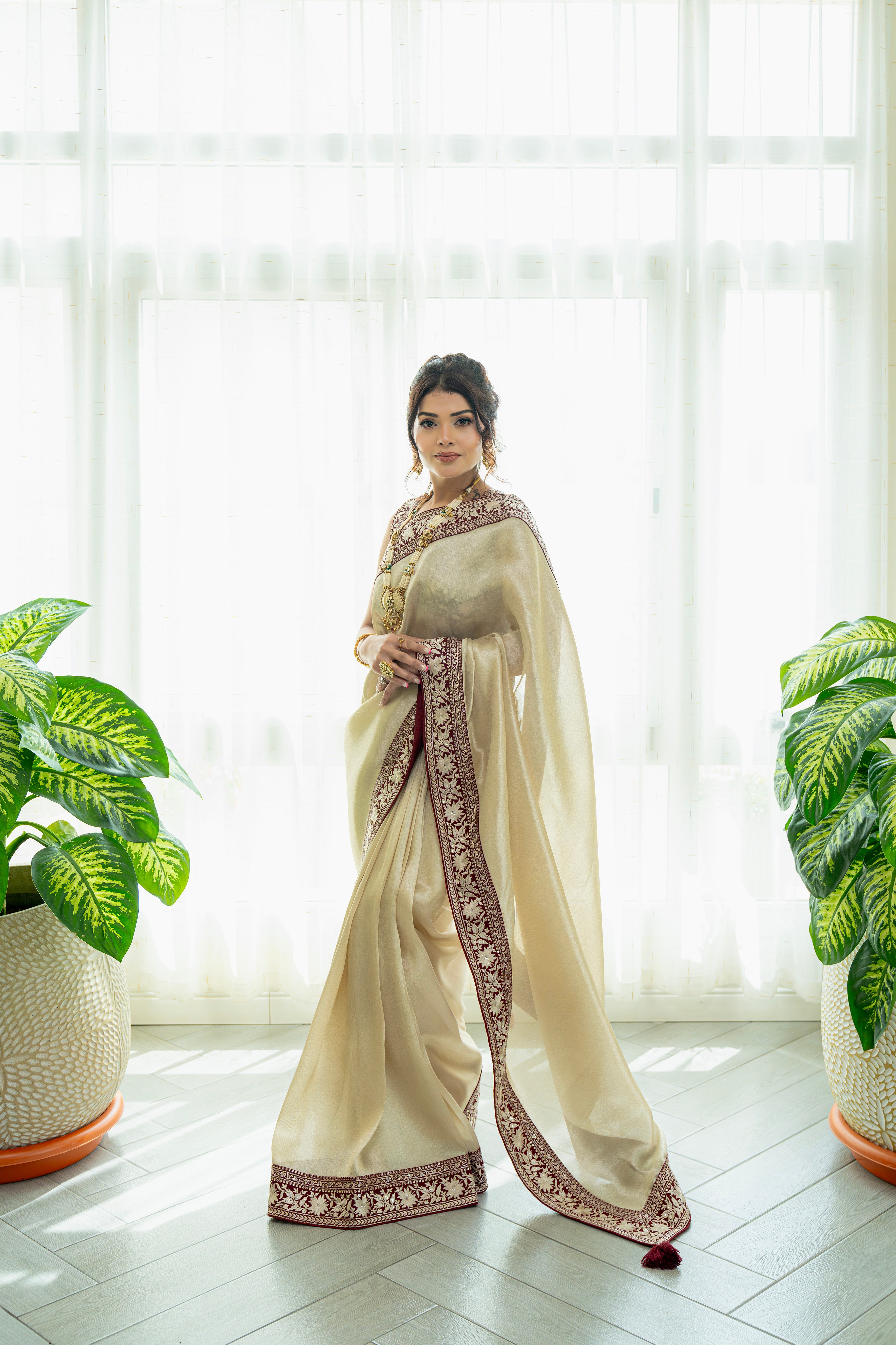 Woman wearing an elegant cream saree with a maroon embroidered border, styled indoors, perfect for weddings and festive occasions.
