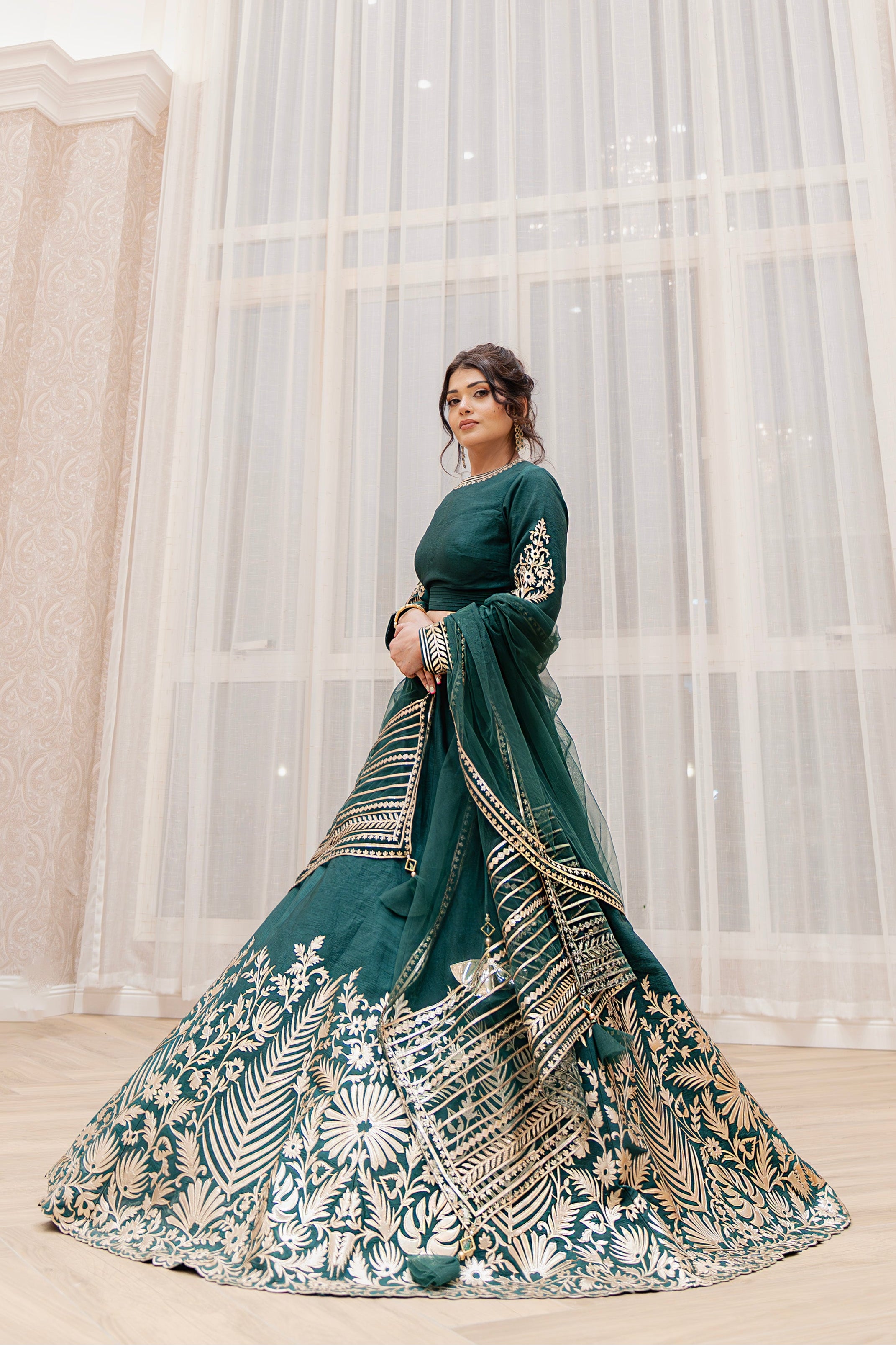 Woman in a green and gold traditional outfit standing in a room with large windows.