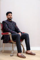Man seated on an ornate chair wearing a black Indo-Western sherwani jacket with red shimmer floral jacquard over a black kurta and trousers, accented with a red pocket square, against a light wallpapered background.