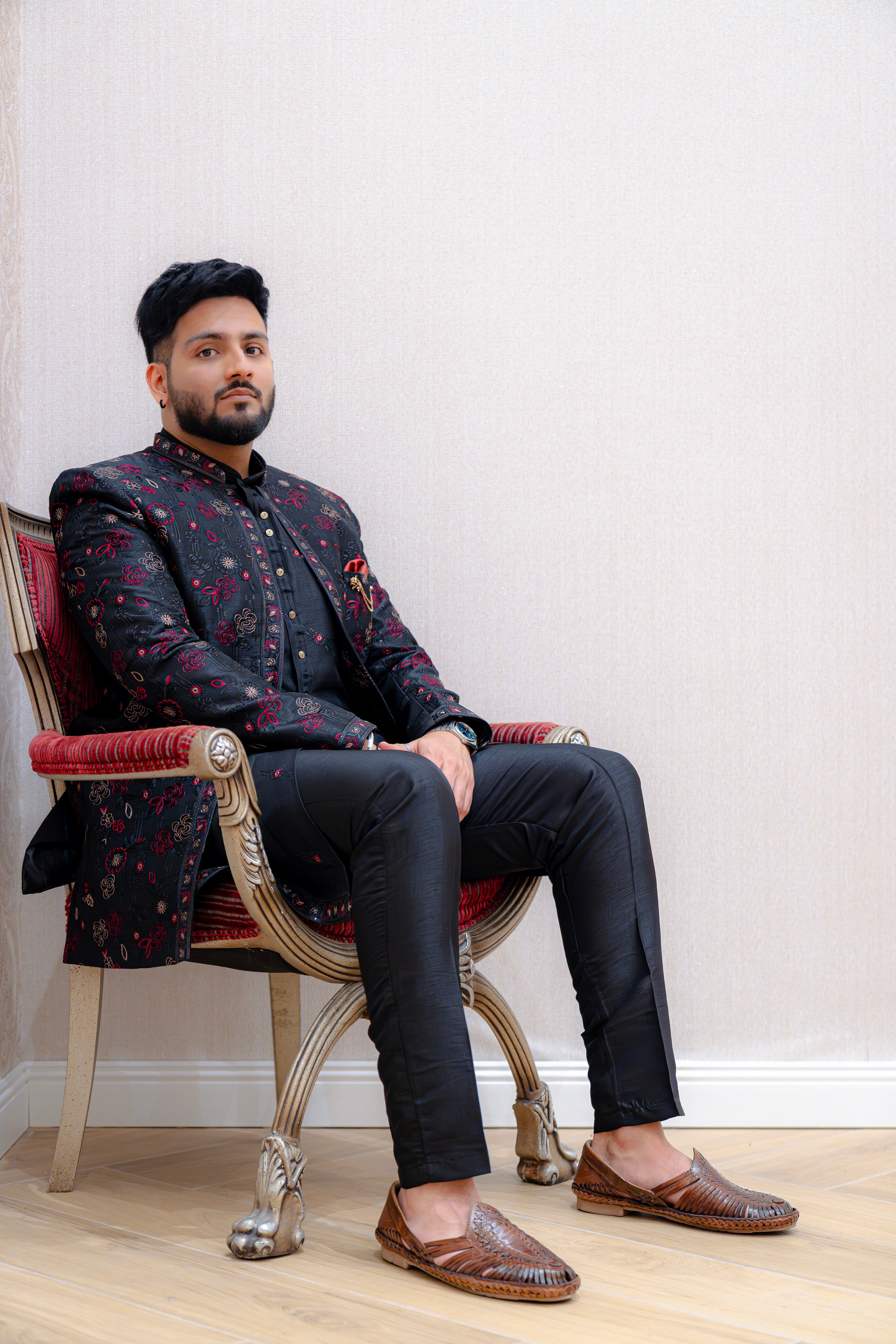 Man seated on an ornate chair wearing a black Indo-Western sherwani jacket with red shimmer floral jacquard over a black kurta and trousers, accented with a red pocket square, against a light wallpapered background.