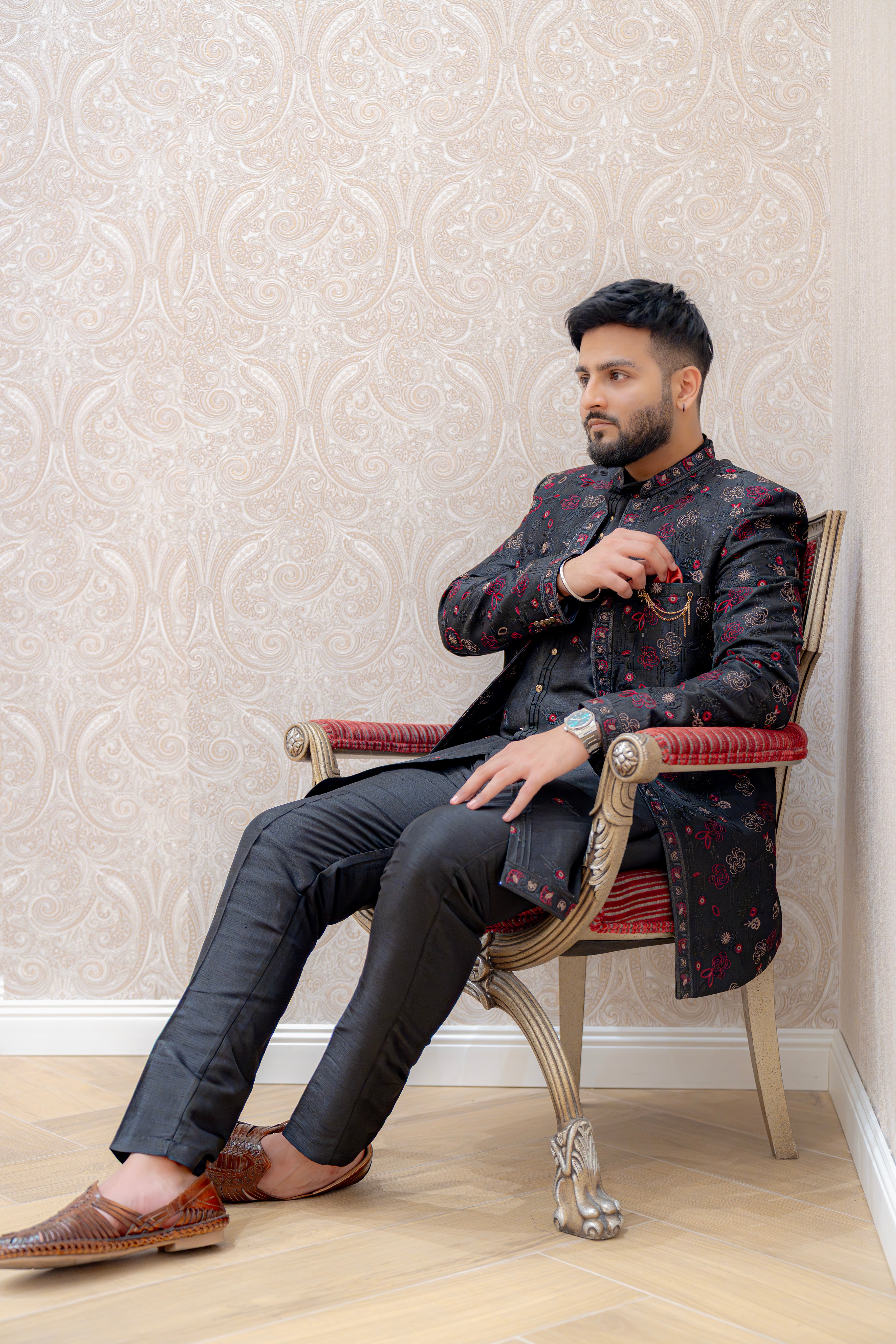 Man in a patterned jacket sitting on a chair against a decorative wall.