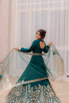 Woman in an emerald traditional outfit with gold embroidery, standing indoors with curtains in the background.