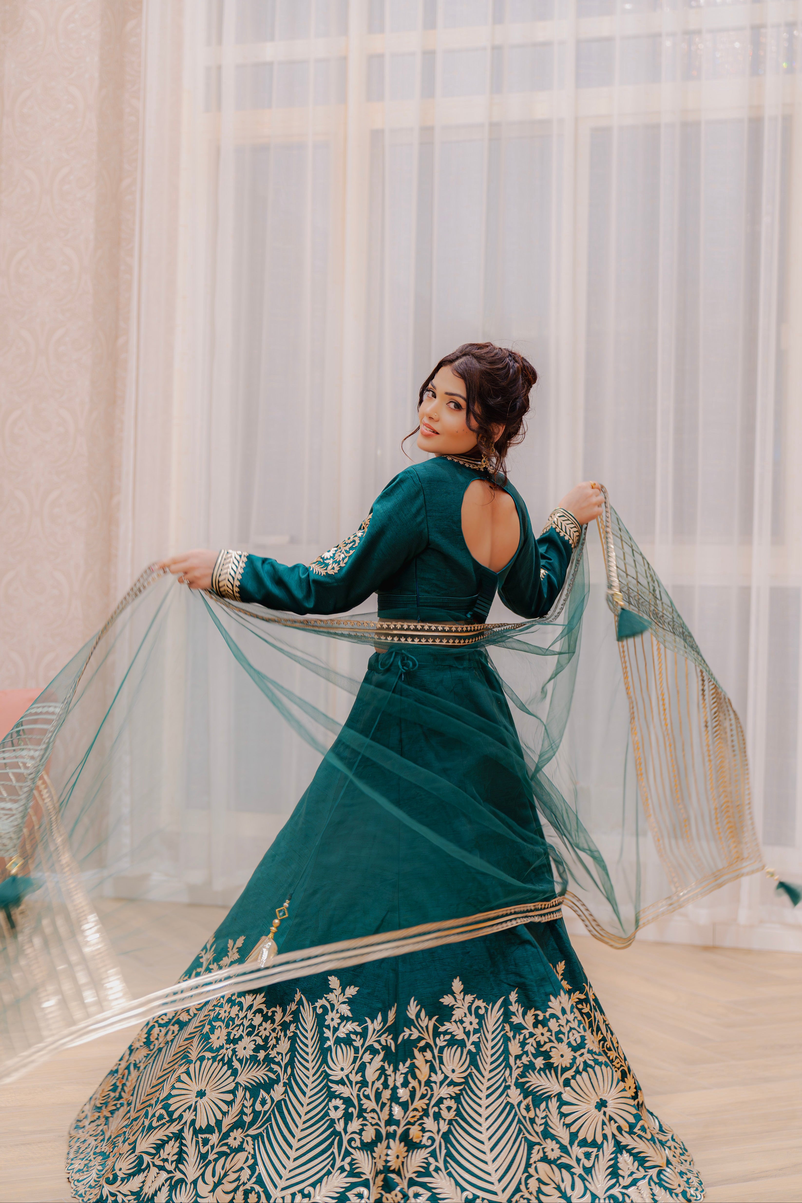 Woman in an emerald traditional outfit with gold embroidery, standing indoors with curtains in the background.