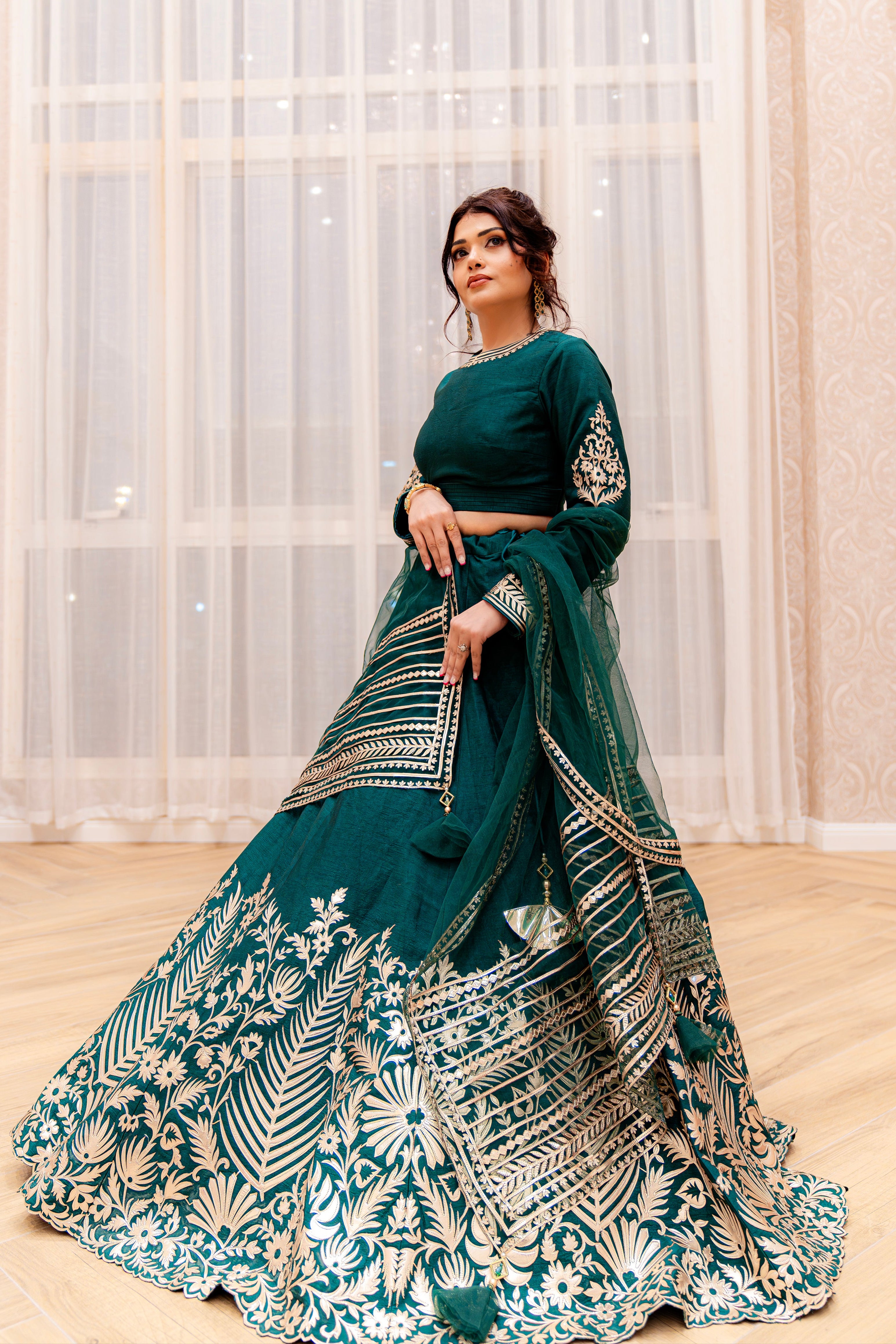 Woman wearing an emerald green lehenga set with gold leather appliqué detailing, full-sleeve blouse and matching net dupatta, standing indoors against sheer curtains.