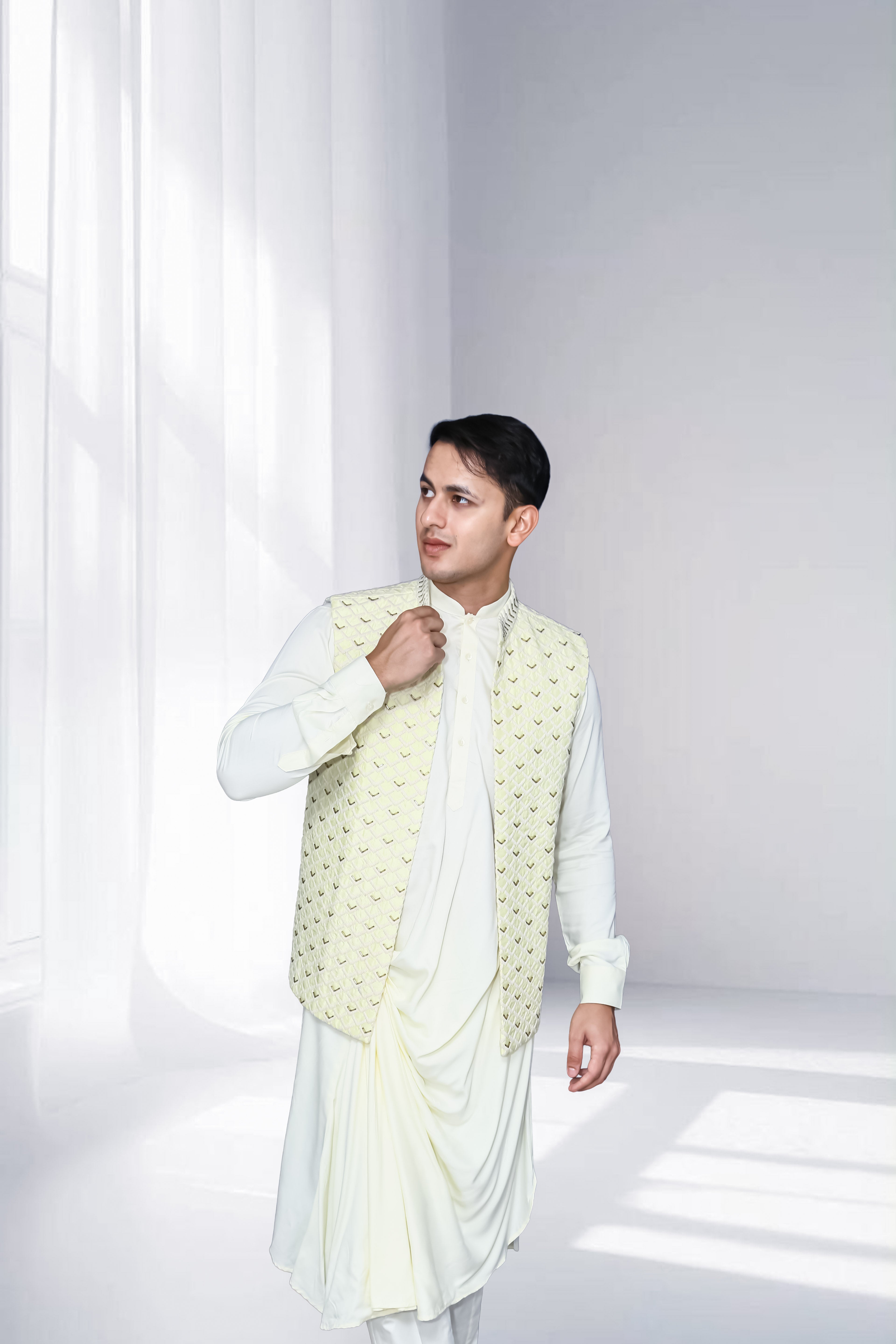 Man in an ivory indo-western kurta with draped detailing, styled with a cream embroidered nehru jacket, standing against a soft-lit minimal background.