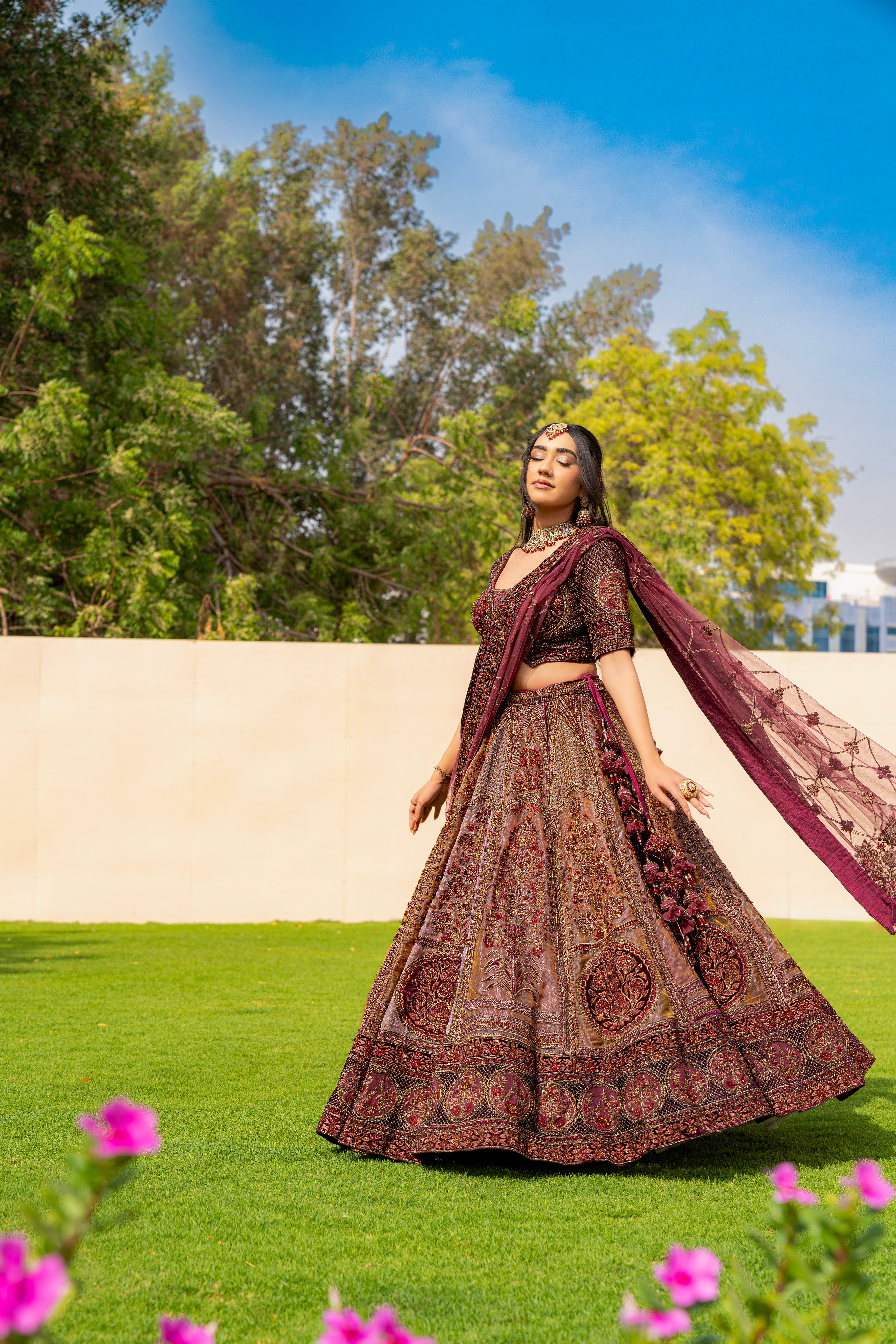 Woman wearing a maroon and brown embroidered bridal lehenga with matching dupatta, standing outdoors, ideal for weddings and receptions.