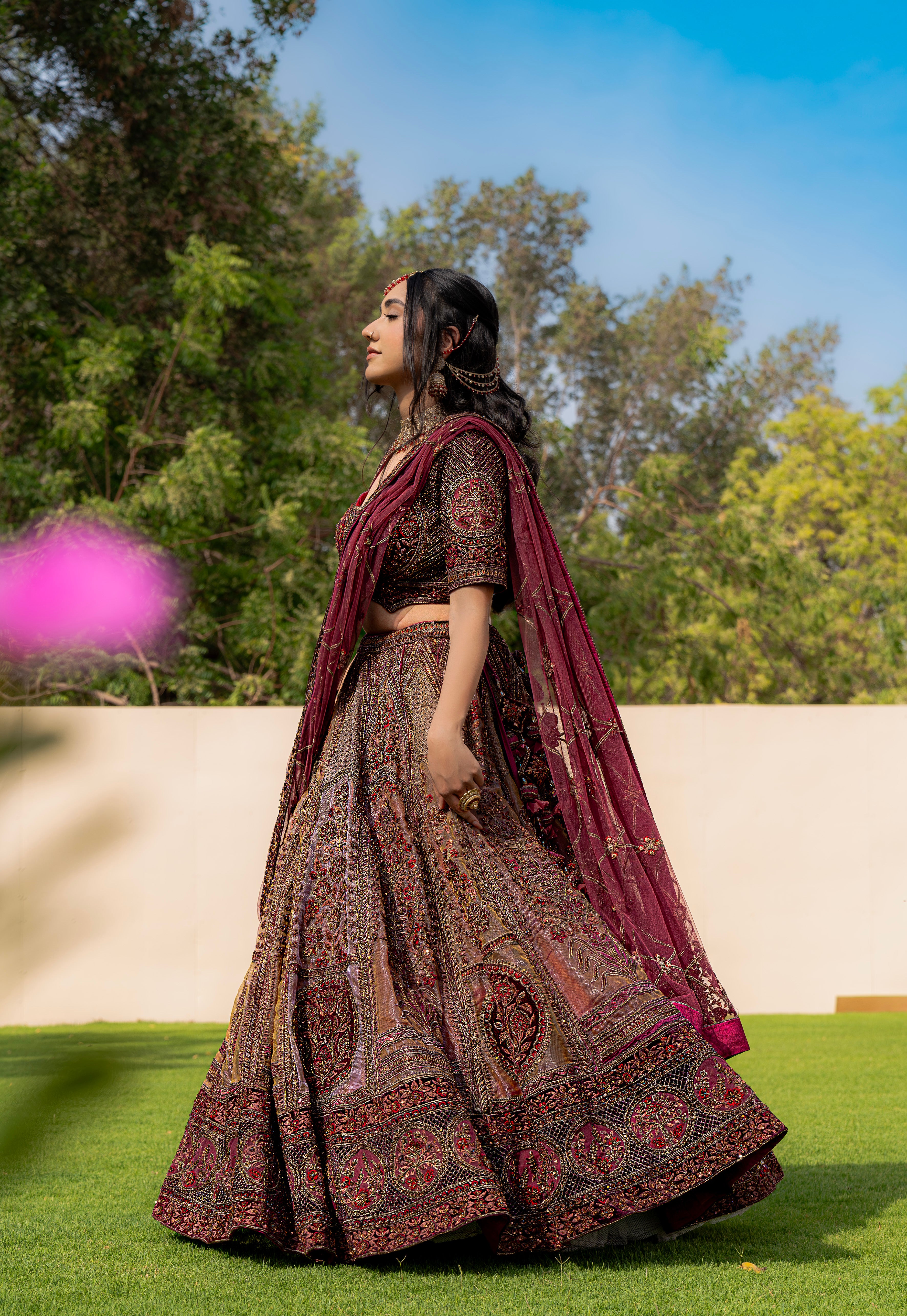 Woman wearing a maroon and brown embroidered bridal lehenga with matching dupatta, standing outdoors, ideal for weddings and receptions.