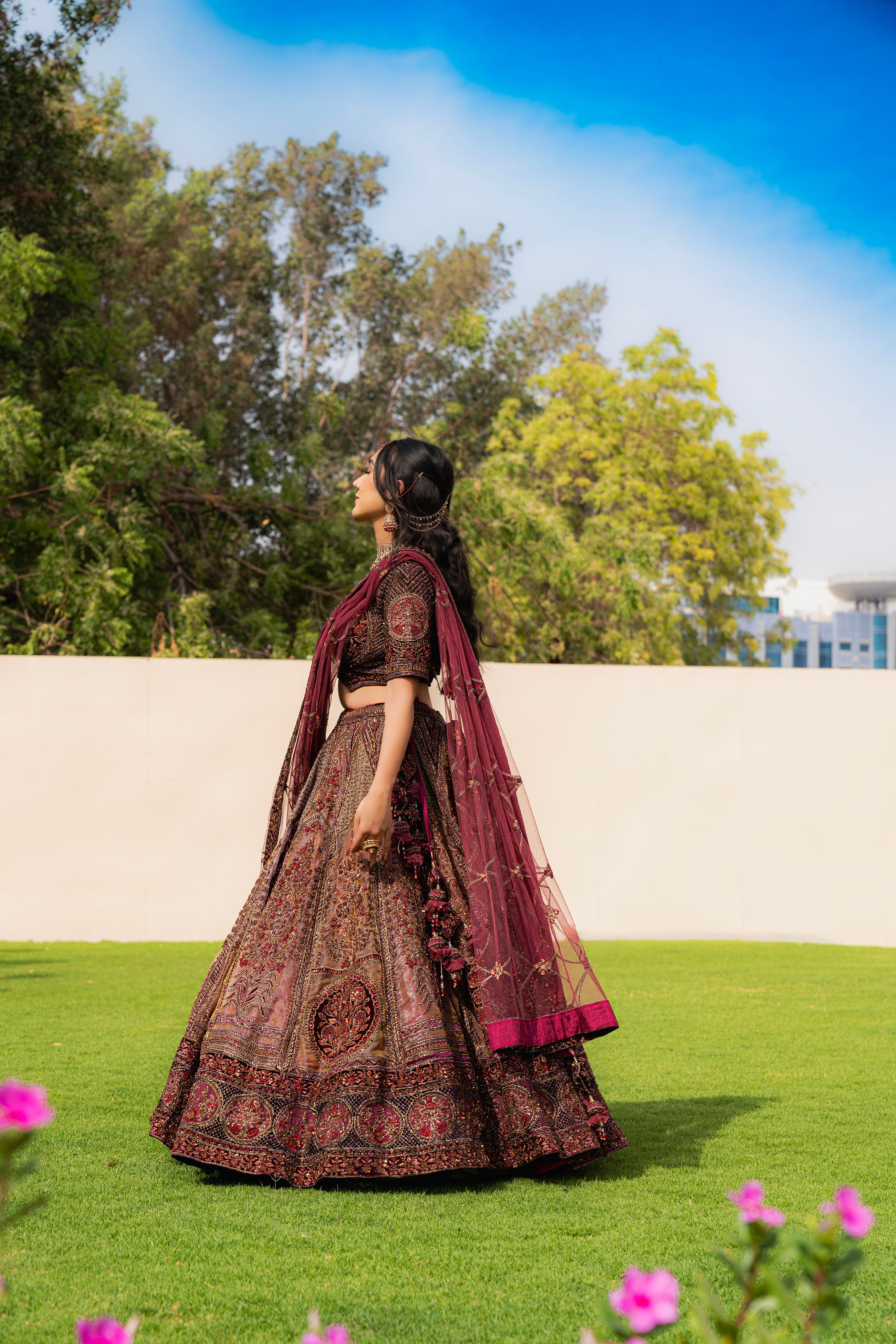 Woman wearing a maroon and brown embroidered bridal lehenga with matching dupatta, standing outdoors, ideal for weddings and receptions.