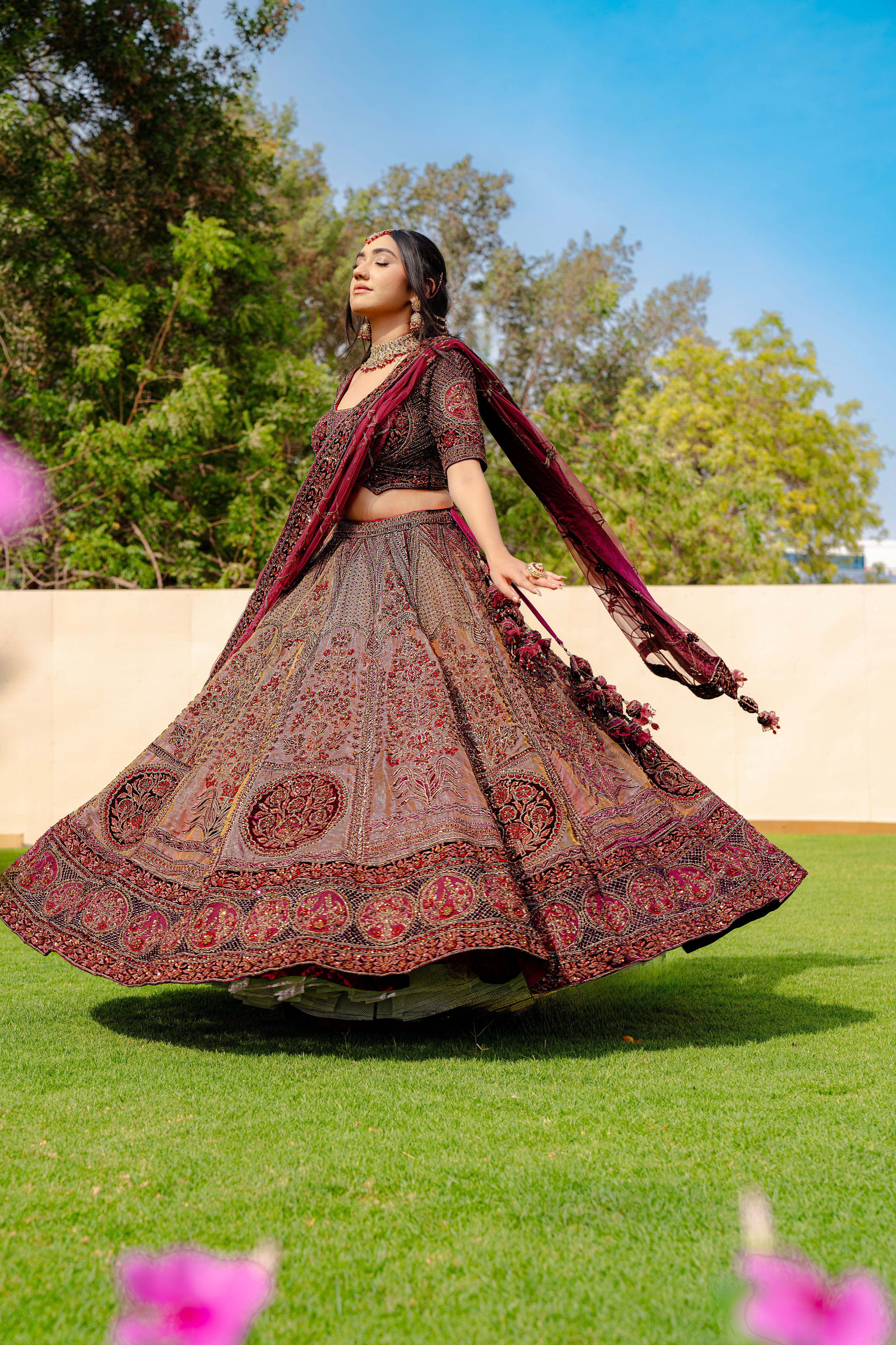 Woman wearing a maroon and brown embroidered bridal lehenga with matching dupatta, standing outdoors, ideal for weddings and receptions.