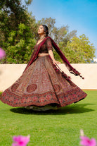 Woman wearing a maroon and brown embroidered bridal lehenga with matching dupatta, standing outdoors, ideal for weddings and receptions.
