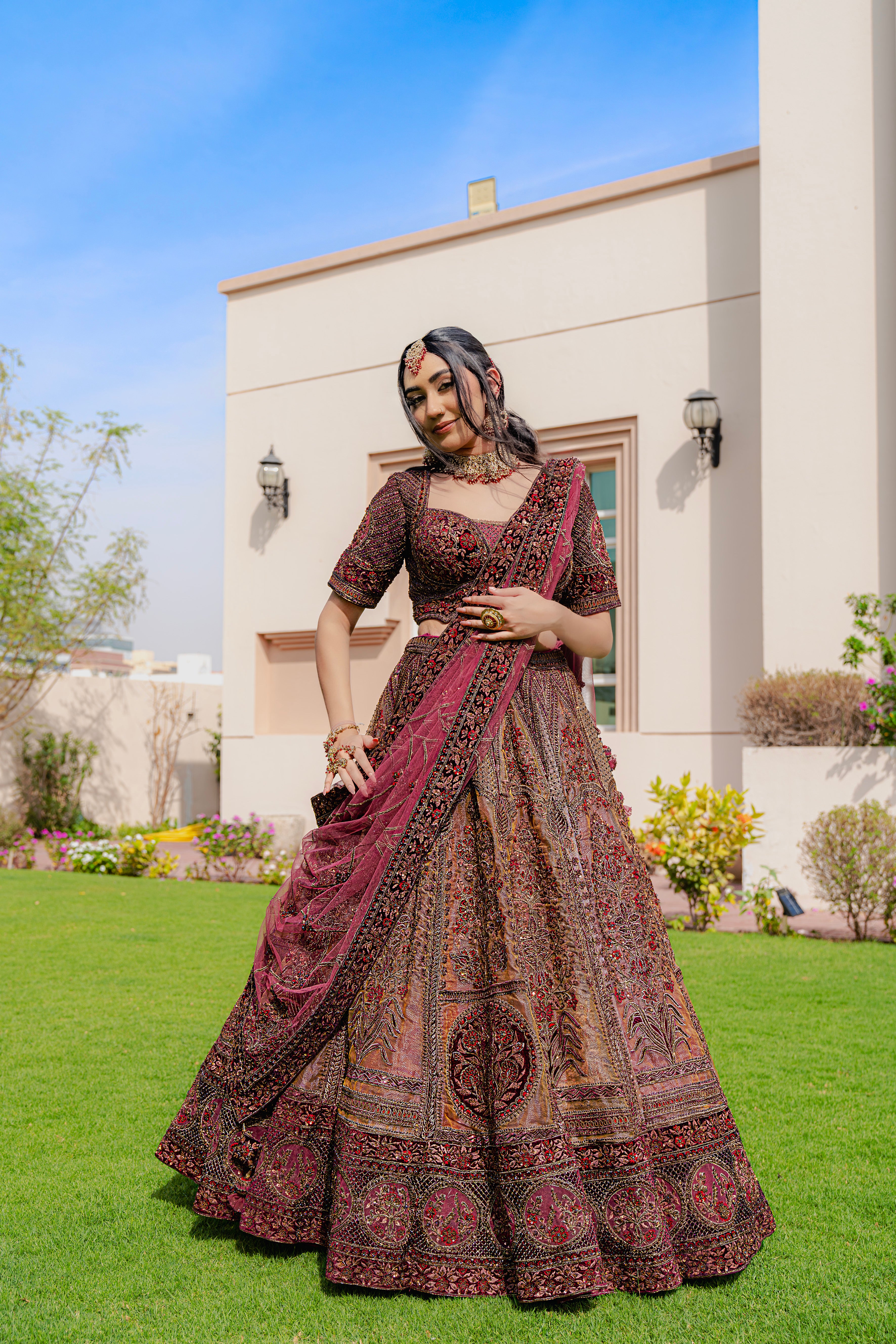 Woman wearing a maroon and brown embroidered bridal lehenga with matching dupatta, standing outdoors, ideal for weddings and receptions.