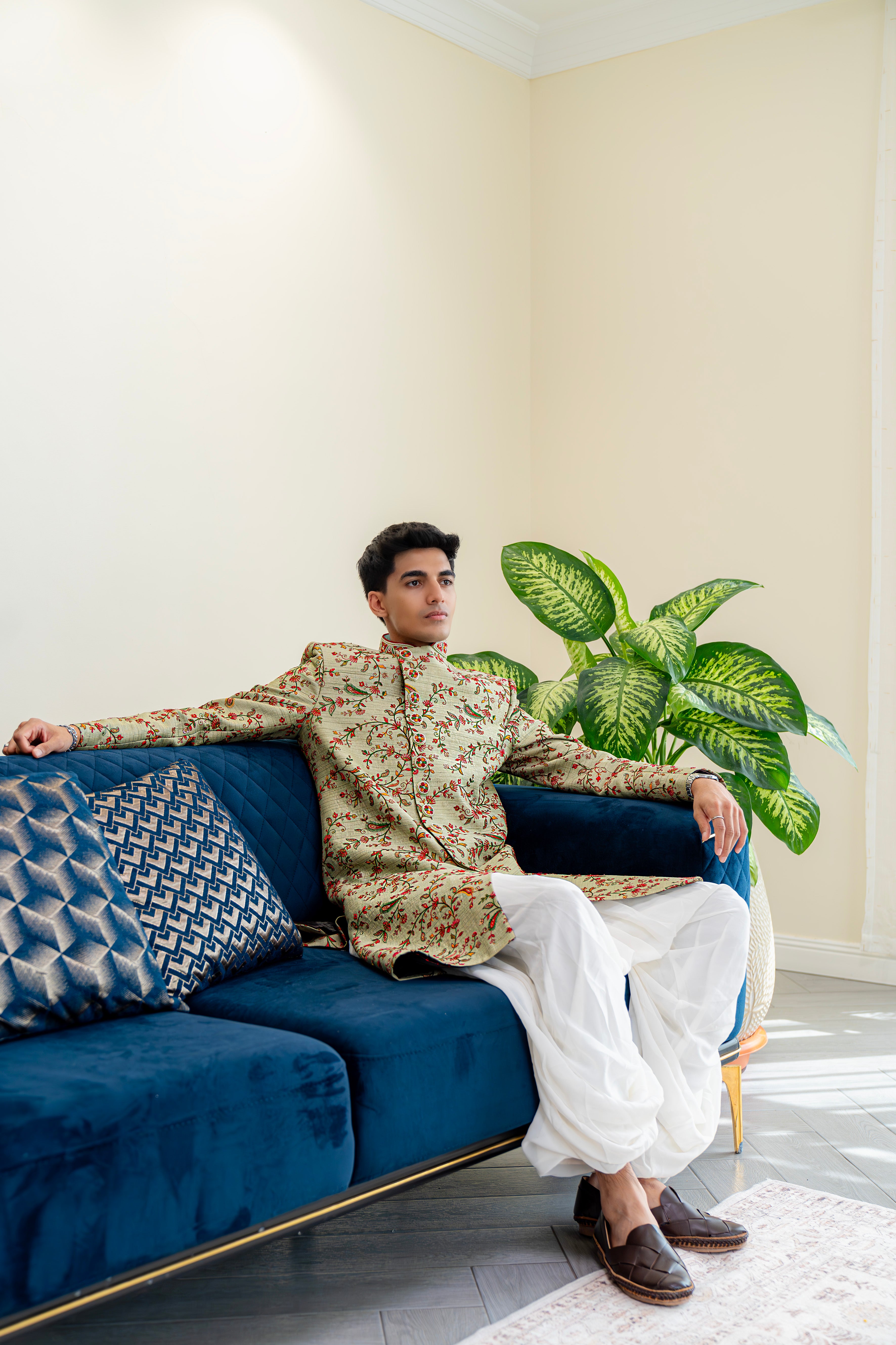 Man wearing an olive green floral embroidered sherwani paired with ivory dhoti pants, styled indoors, ideal for weddings and festive occasions.