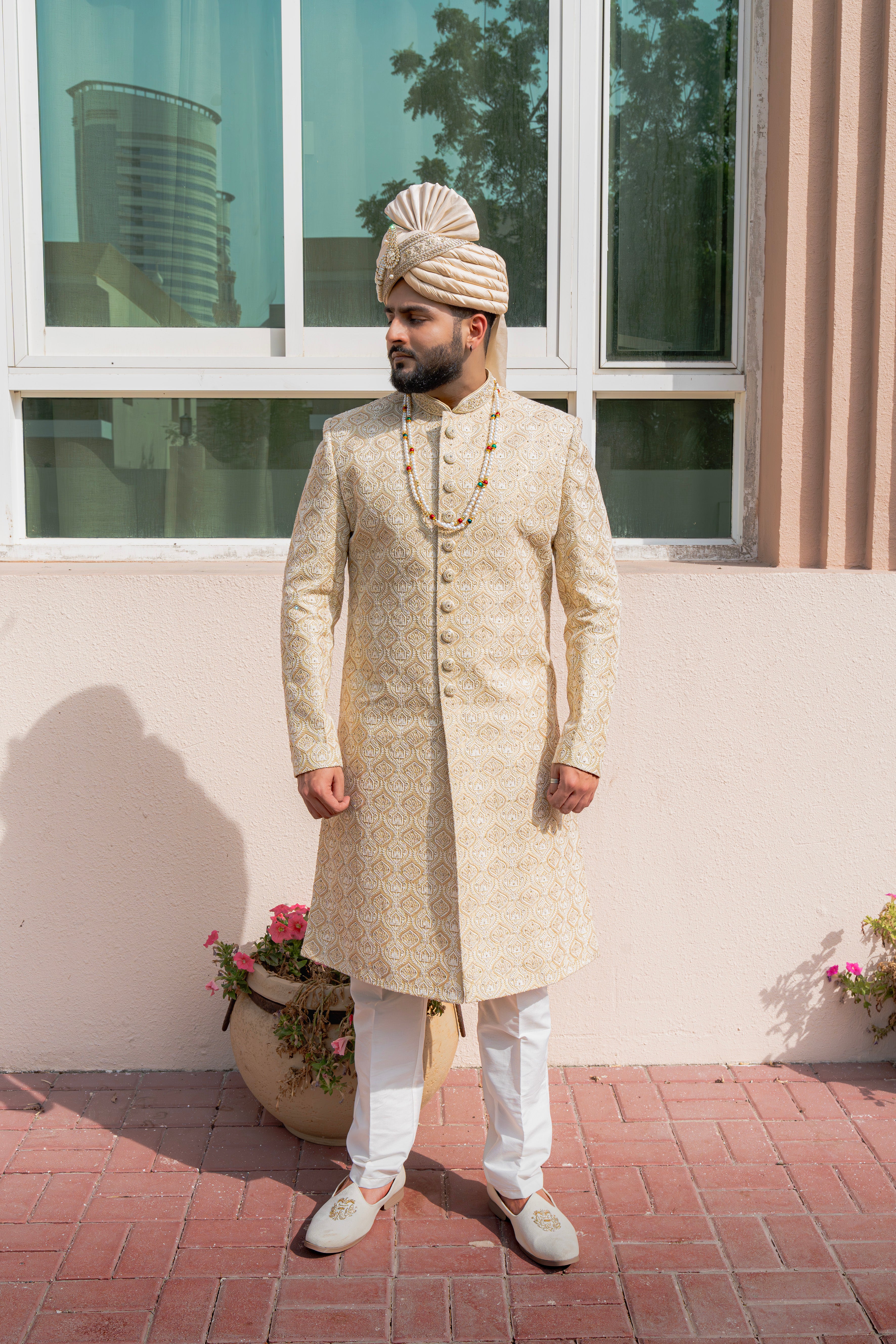 Man in traditional pale yellow sherwani with a safa standing outdoors against a building.